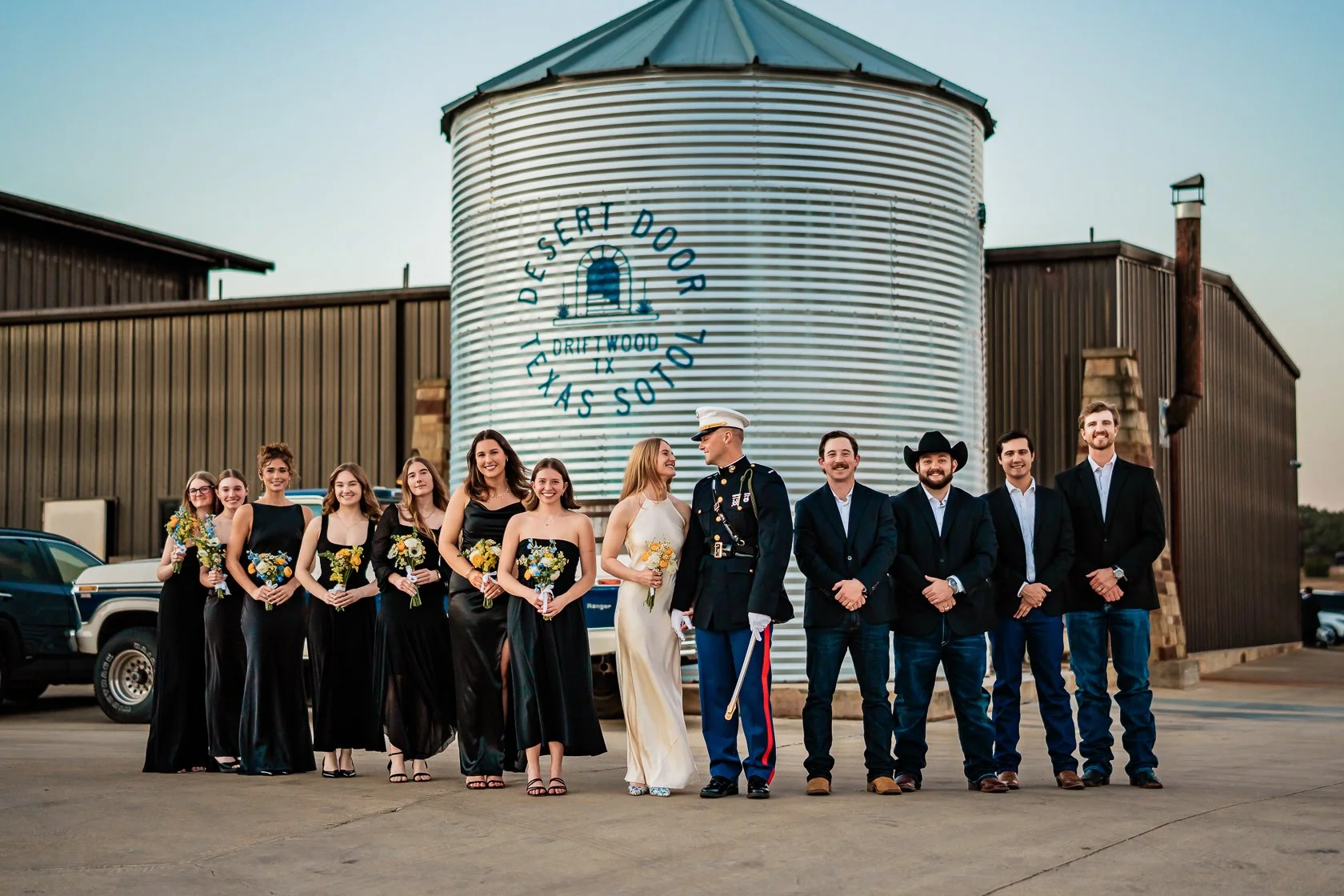 Group of people dressed in formal attire, including women in black dresses holding bouquets and men in suits, standing outside near a large metal grain silo with a sign that reads 'Descert Door Shall Not Pass, Driett Wood, TX'.