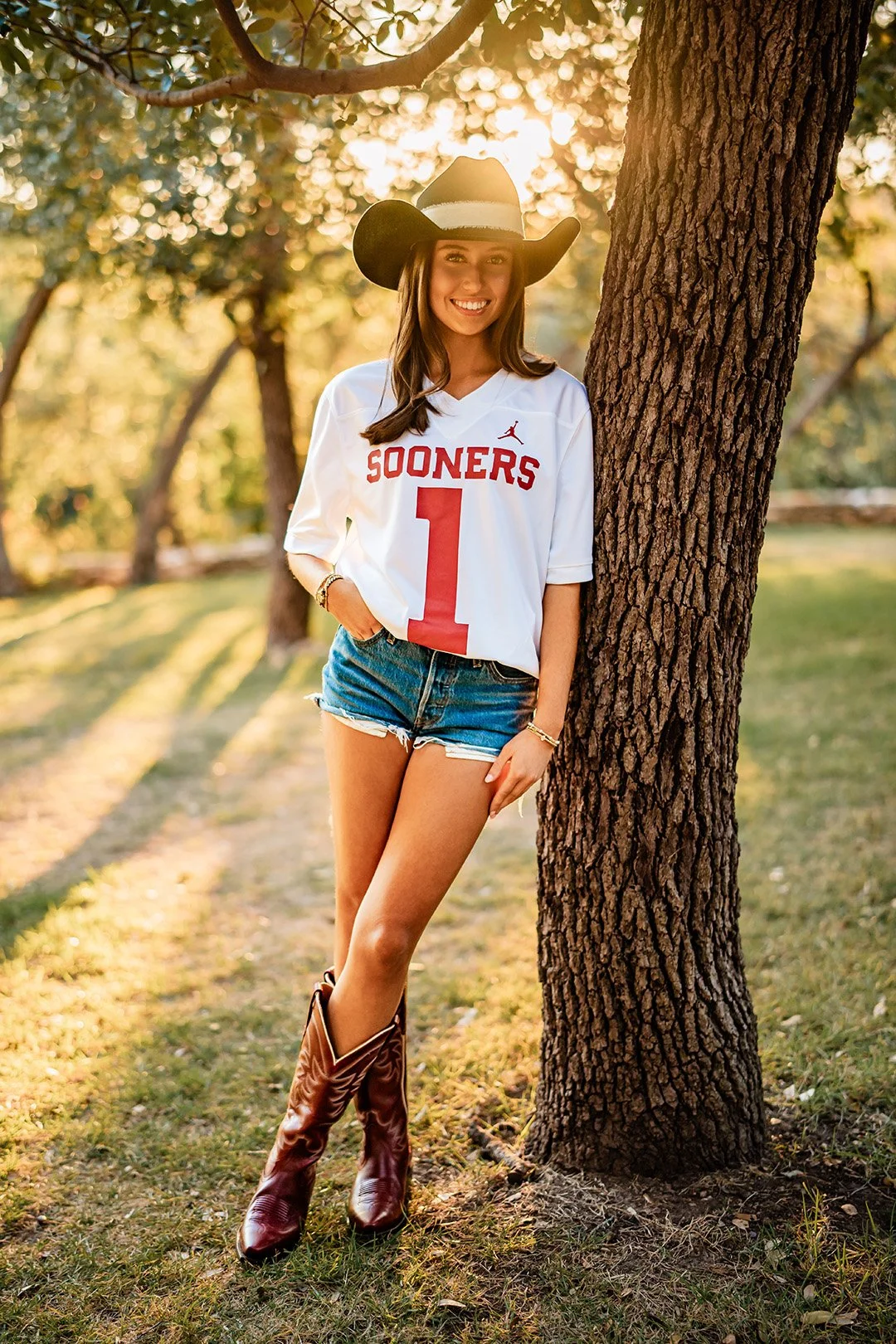 A young woman standing outdoors next to a tree, wearing a white Oklahoma Sooners football jersey, denim shorts, cowboy boots, and a cowboy hat, smiling at the camera during sunset.