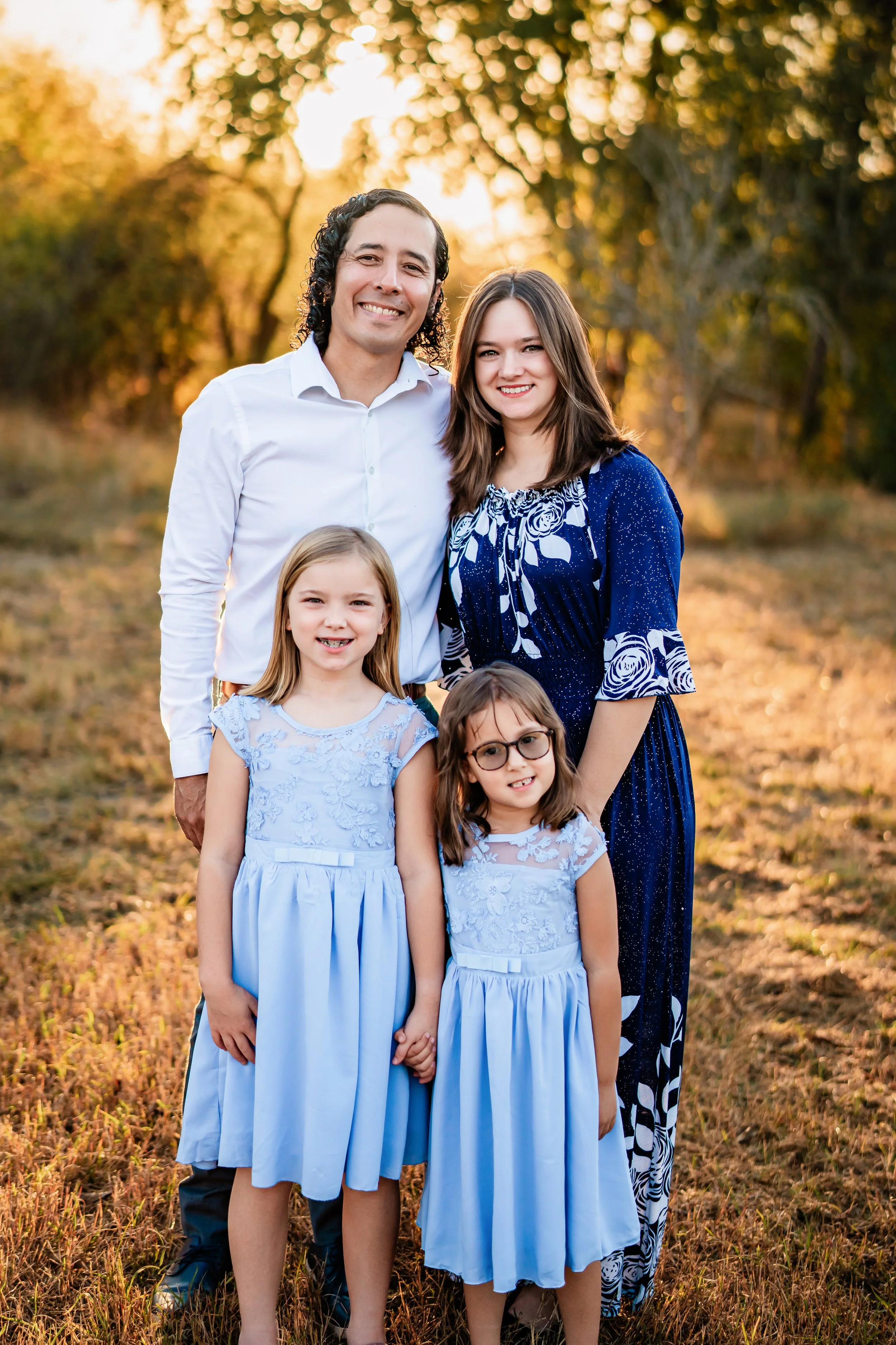 A family of five posing outdoors during sunset. The group includes a man, woman, and three young girls. The man has curly dark hair and is wearing a white shirt. The woman has long brown hair and is wearing a blue dress with white floral patterns. Th