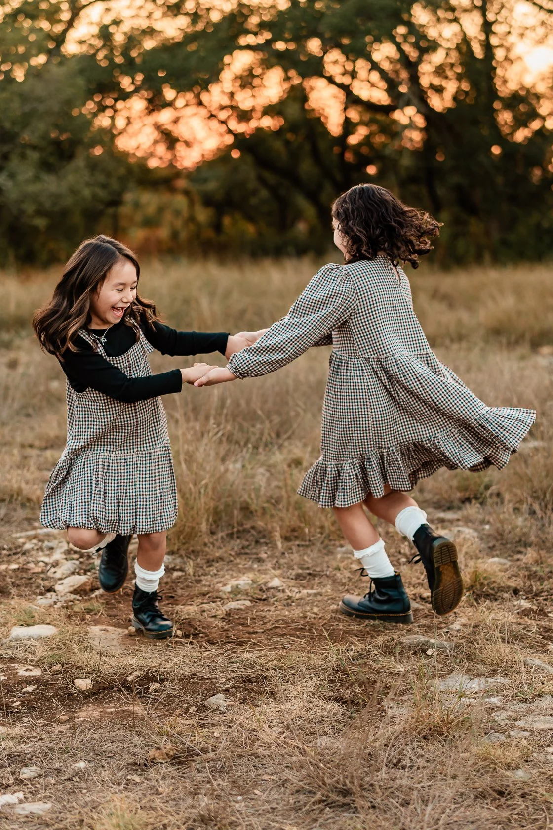 Two young girls in plaid dresses playing and spinning around in a field with tall grass during sunset.