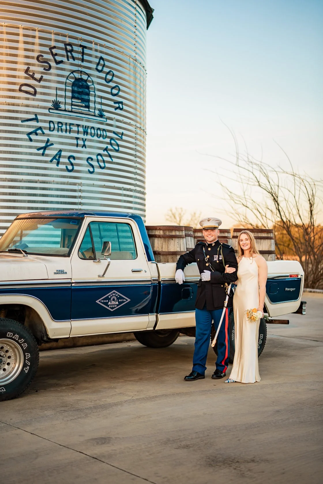 A couple dressed in wedding attire standing next to a pickup truck with a large water tower in the background that reads 'Desert Door Tejas Sott, Driftwood TX.' The groom is in a military uniform, and the bride is in a white dress holding a small bou