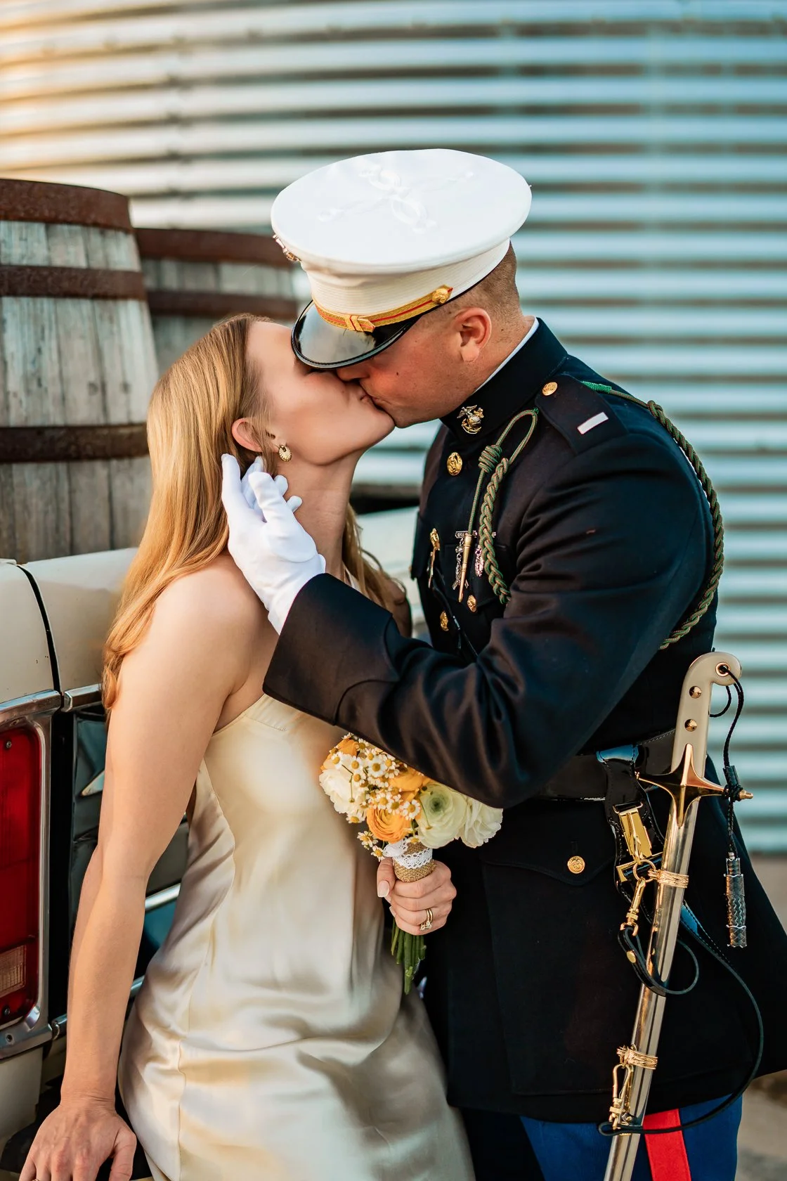 A soldier in uniform kisses a woman in a wedding dress. The woman holds a bouquet of flowers and leans against a vehicle. The soldier wears a white hat and gloves, and has a ceremonial sword attached to his uniform.