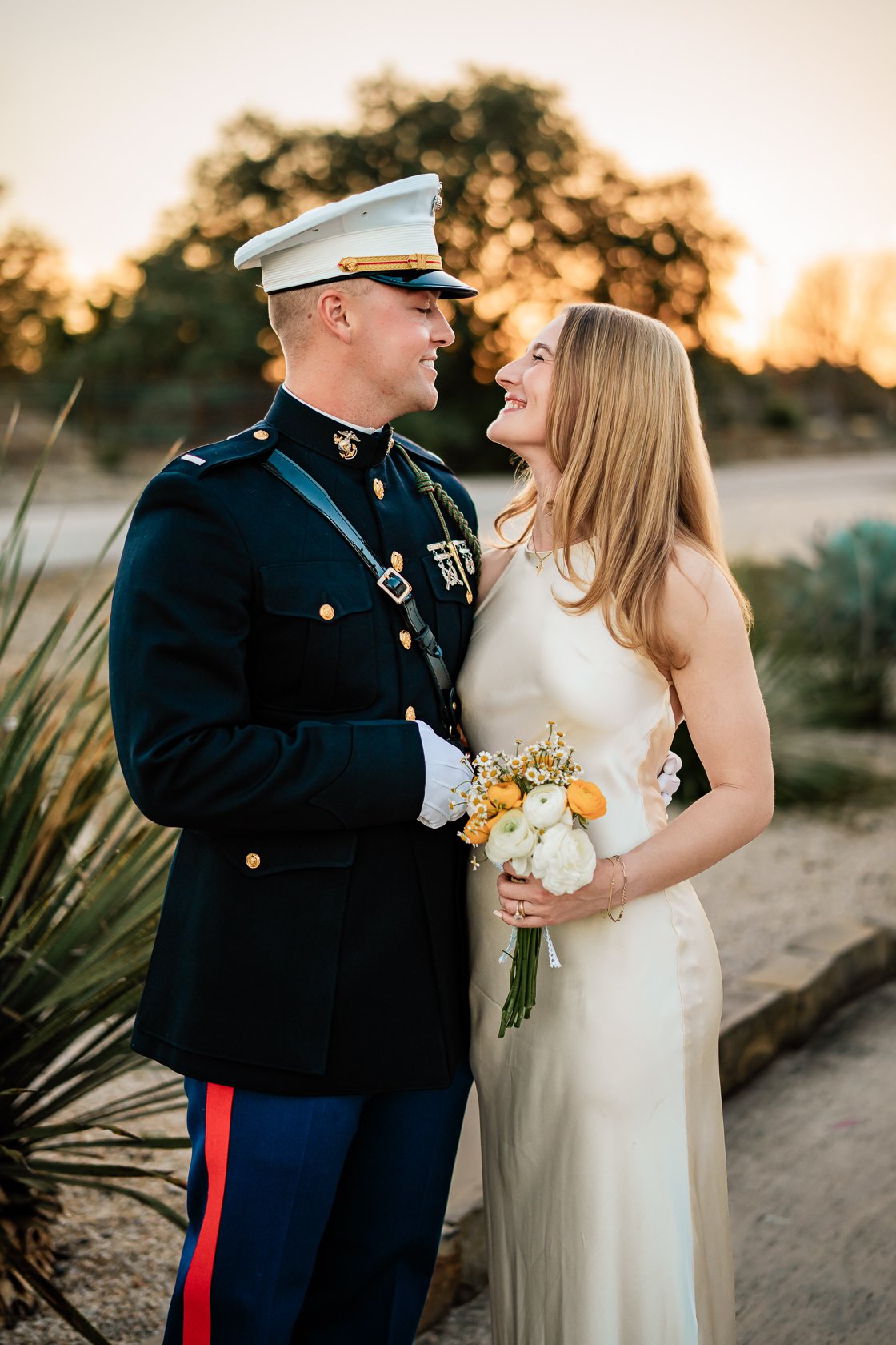 A bride and a groom in uniform stand close, smiling at each other, during their wedding at sunset. The bride holds a bouquet of white and orange flowers.