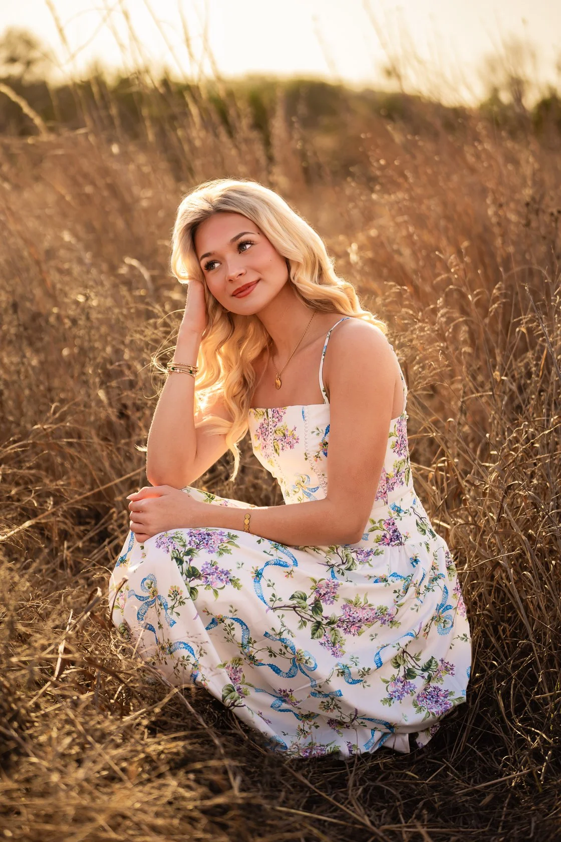 A young woman with long blonde hair sitting in a field of dry grass during sunset, wearing a floral dress and looking thoughtfully to the side.