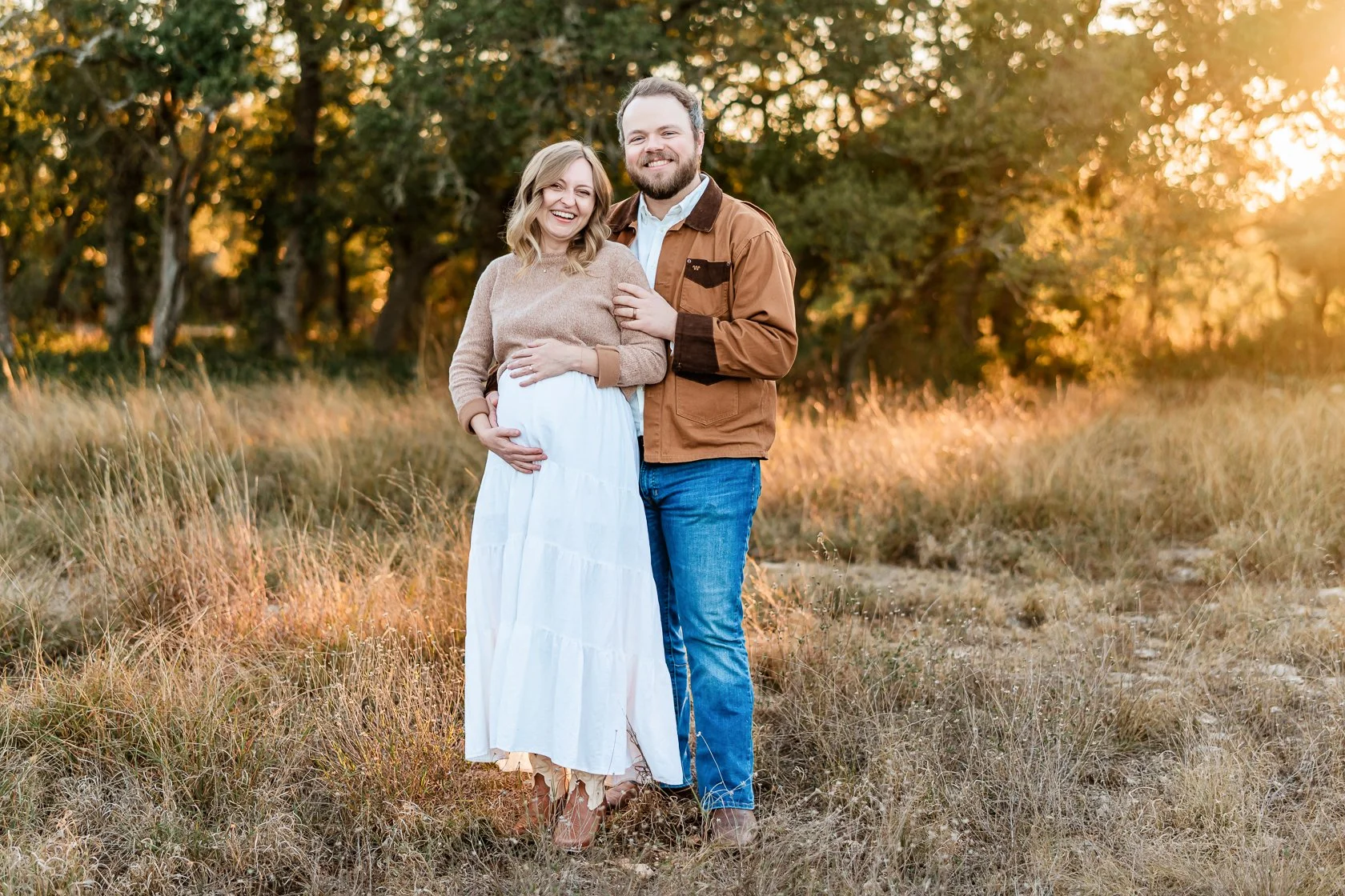 A pregnant woman and her partner standing outdoors in a grassy field during sunset, smiling, with trees in the background.