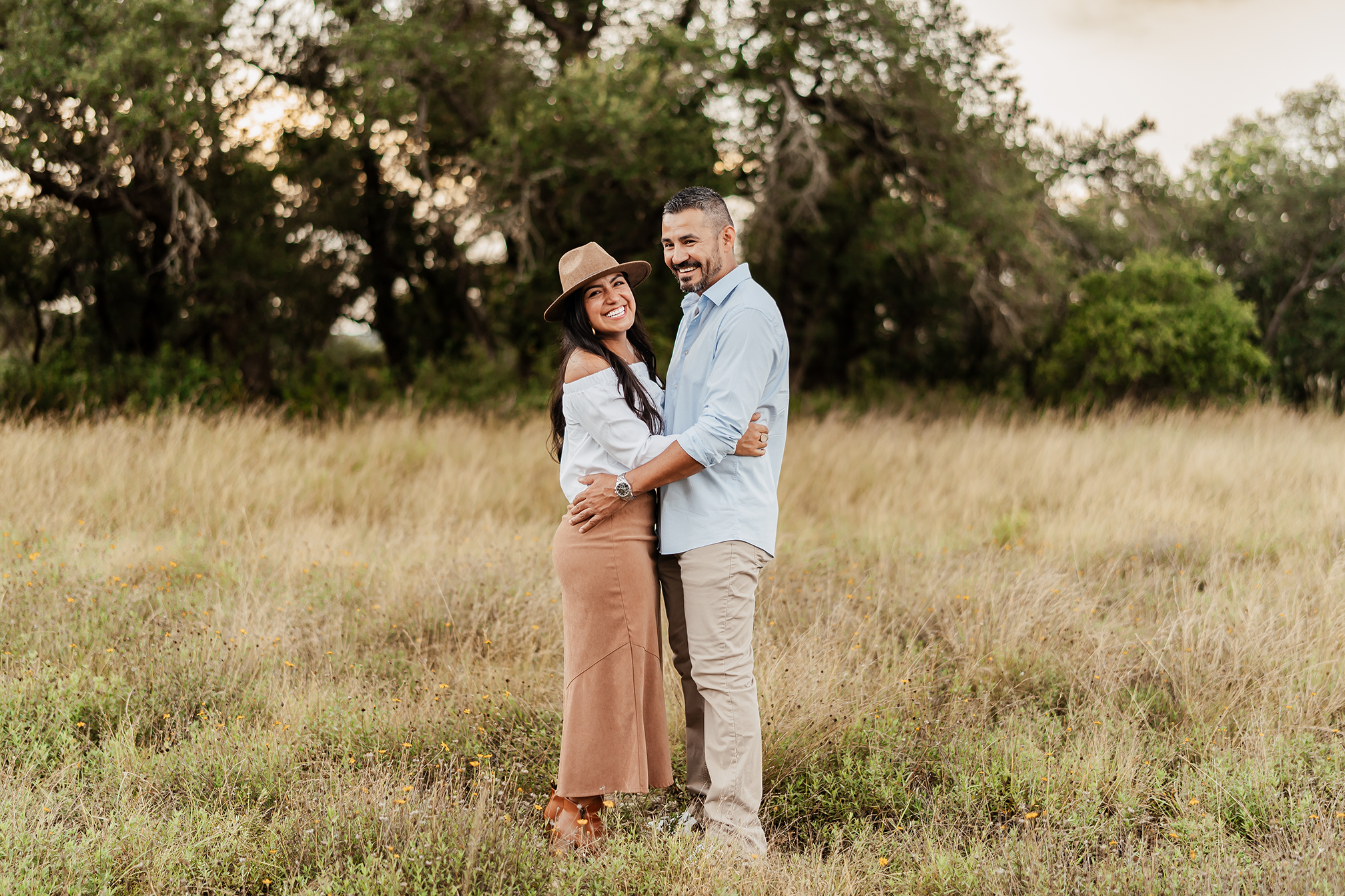A smiling couple standing in a grassy field with trees in the background, embracing each other. The woman wears a hat, white off-shoulder top, and tan skirt, while the man wears a light blue shirt and beige pants.