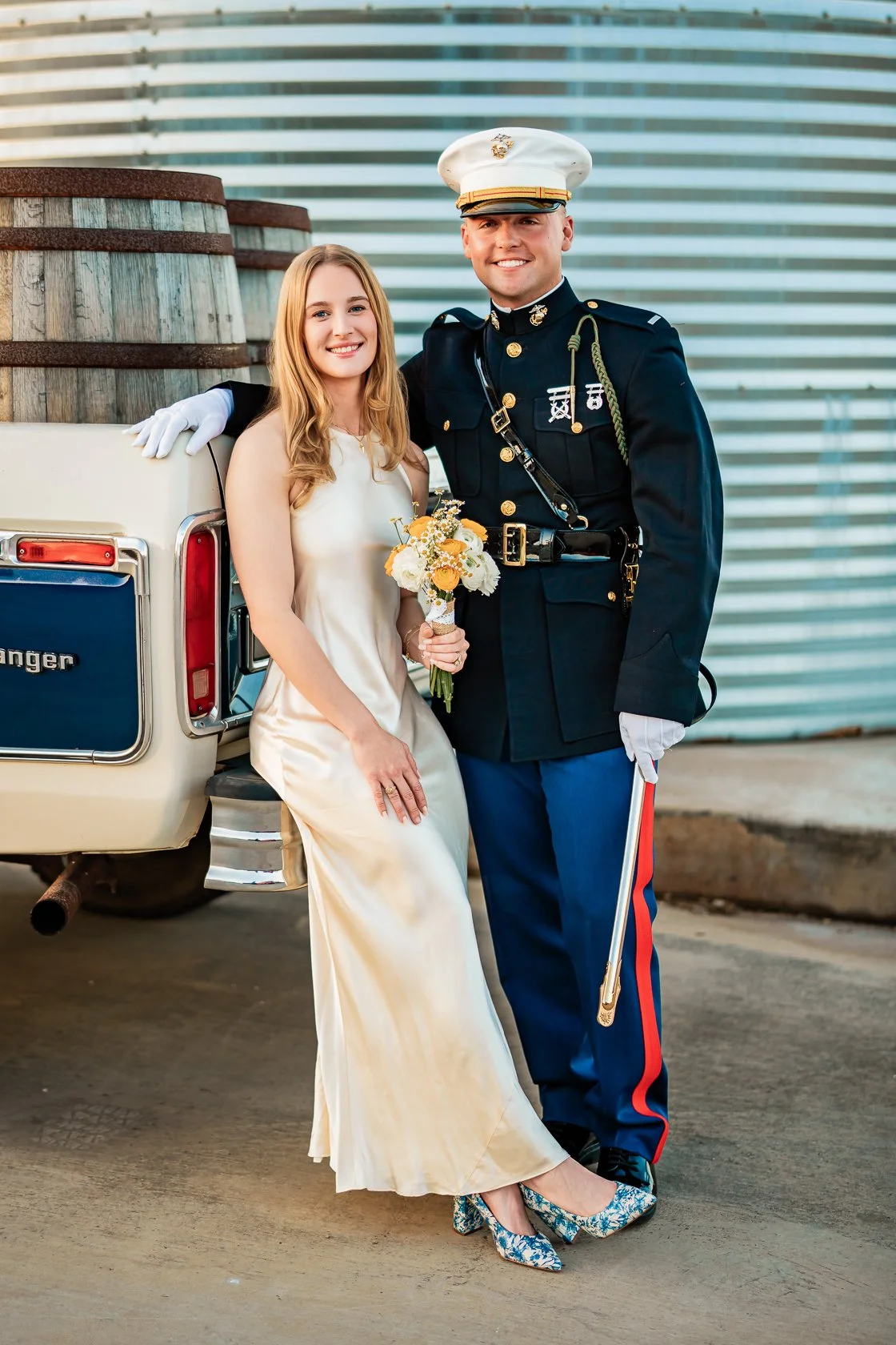 A bride in a white dress holding a bouquet and a groom in a military uniform posing together outdoors next to a vintage car with barrels in the background.