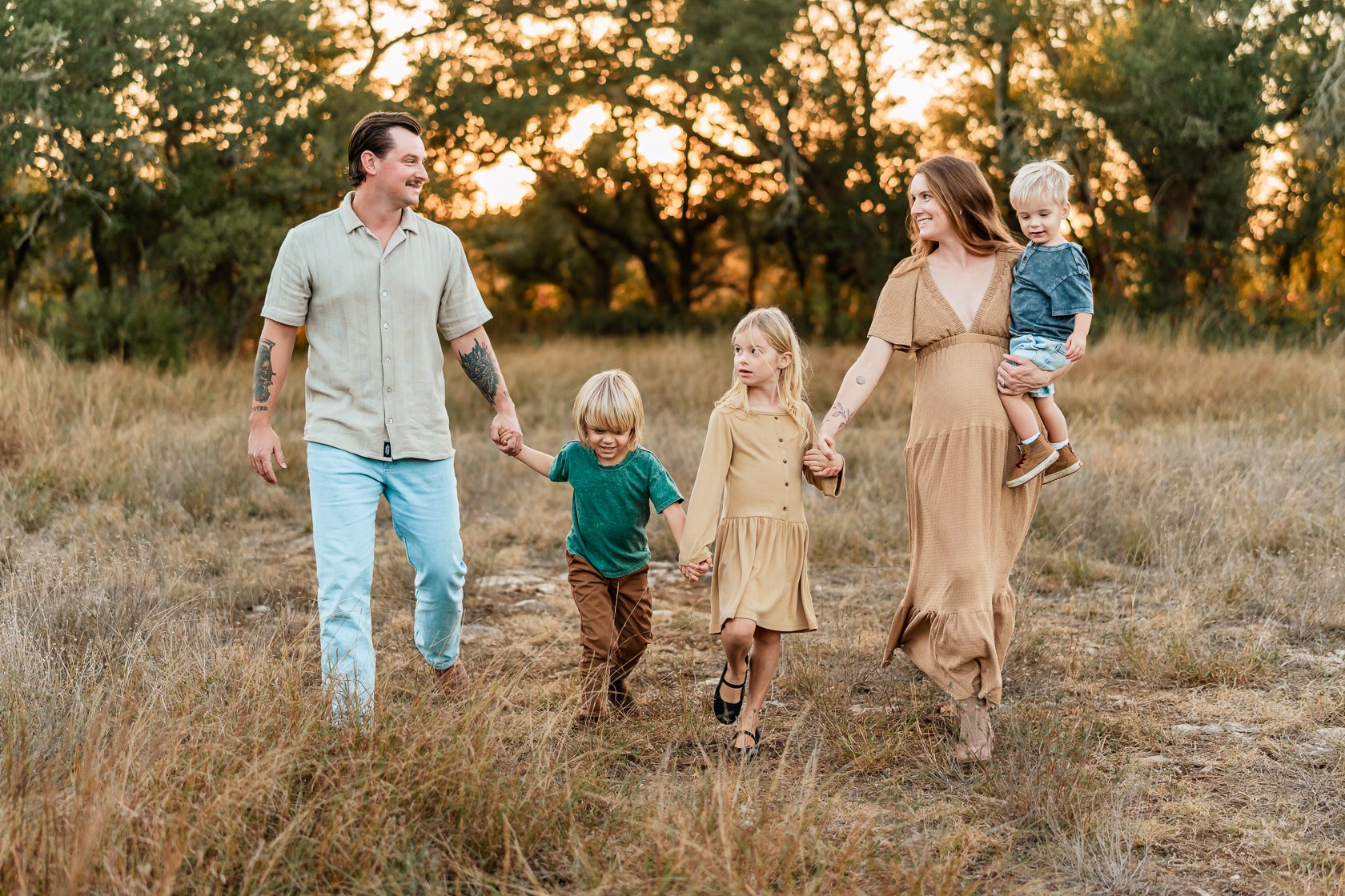 A family of five, including a father, mother, two young girls, and a young boy, walking together hand in hand through a grassy field during sunset.