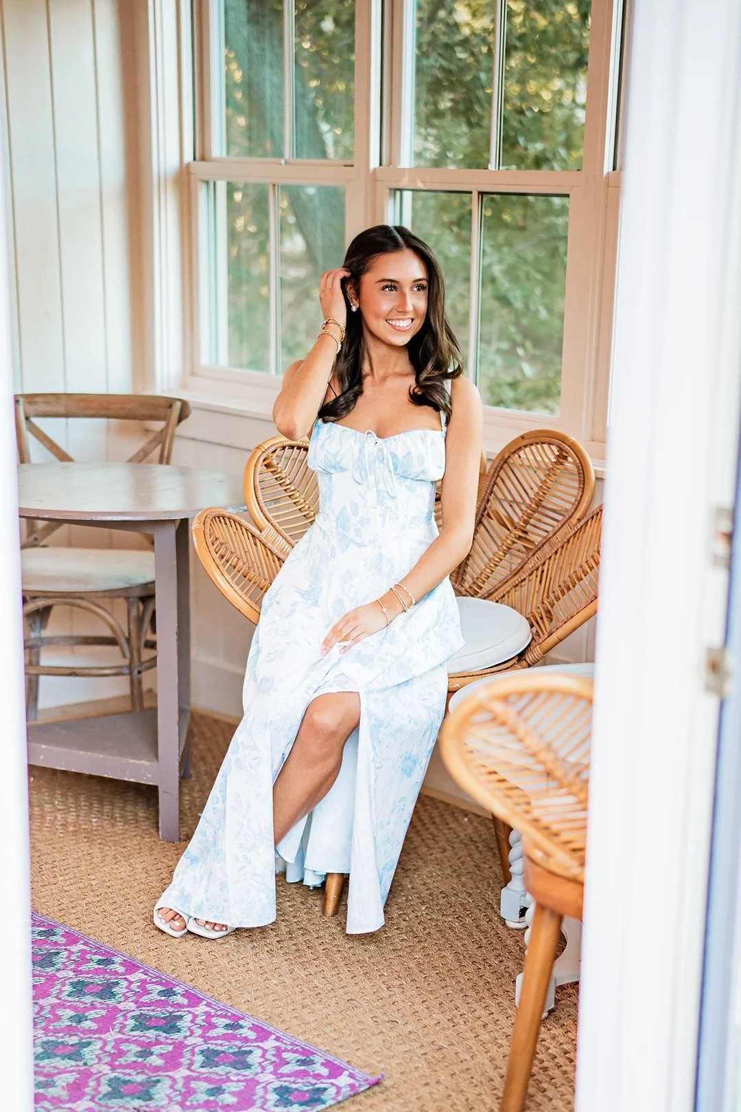 A young woman in a white floral dress sitting on a wicker chair in a sunlit room with large windows and wooden furniture.