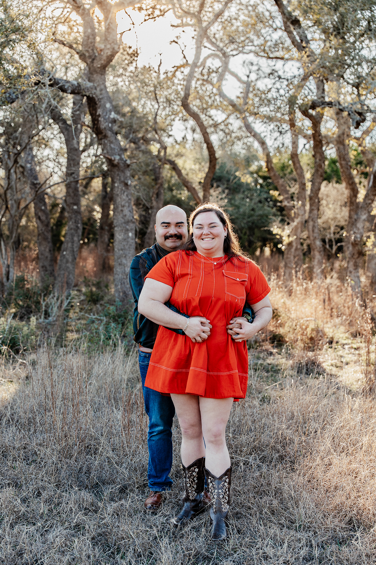 A smiling couple embracing in a sunlit outdoor setting with trees and dry grass, woman wearing a red dress and cowboy boots, man wearing a dark shirt and jeans.