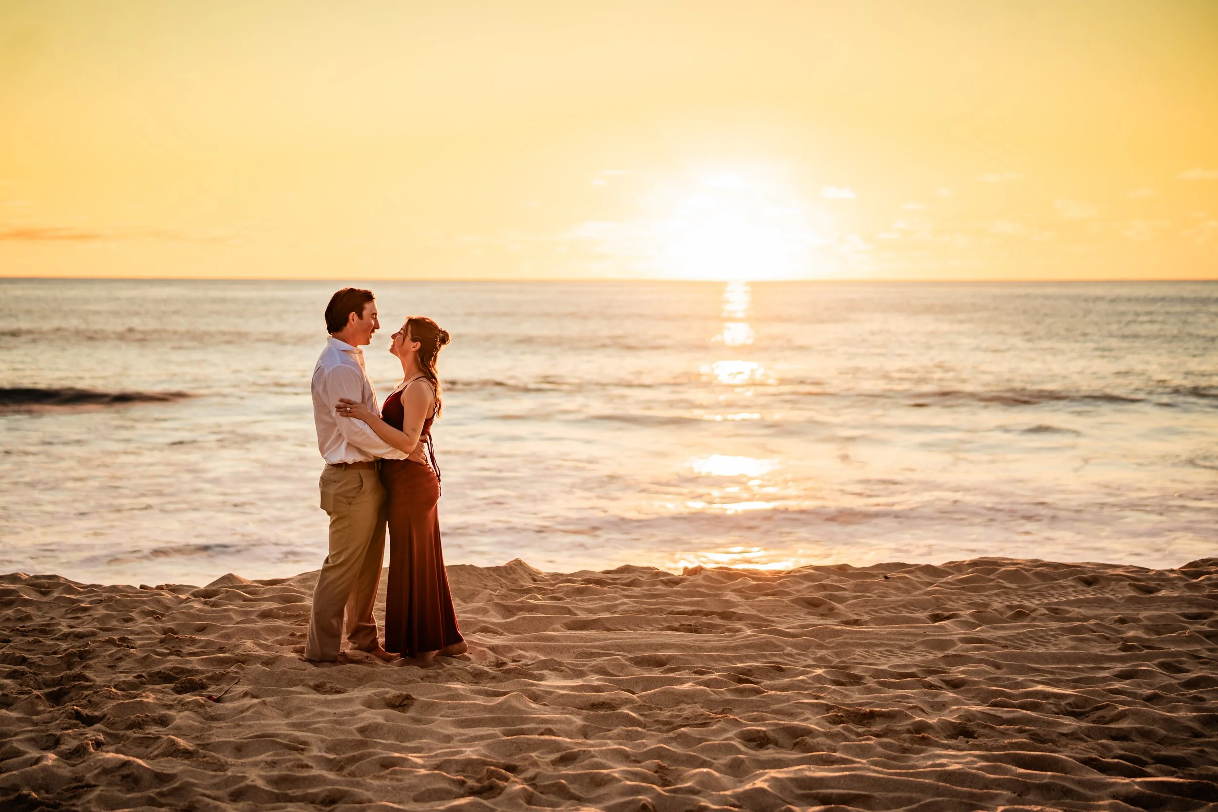 A couple stands close together on a sandy beach at sunset, gazing into each other's eyes with the ocean and setting sun in the background.