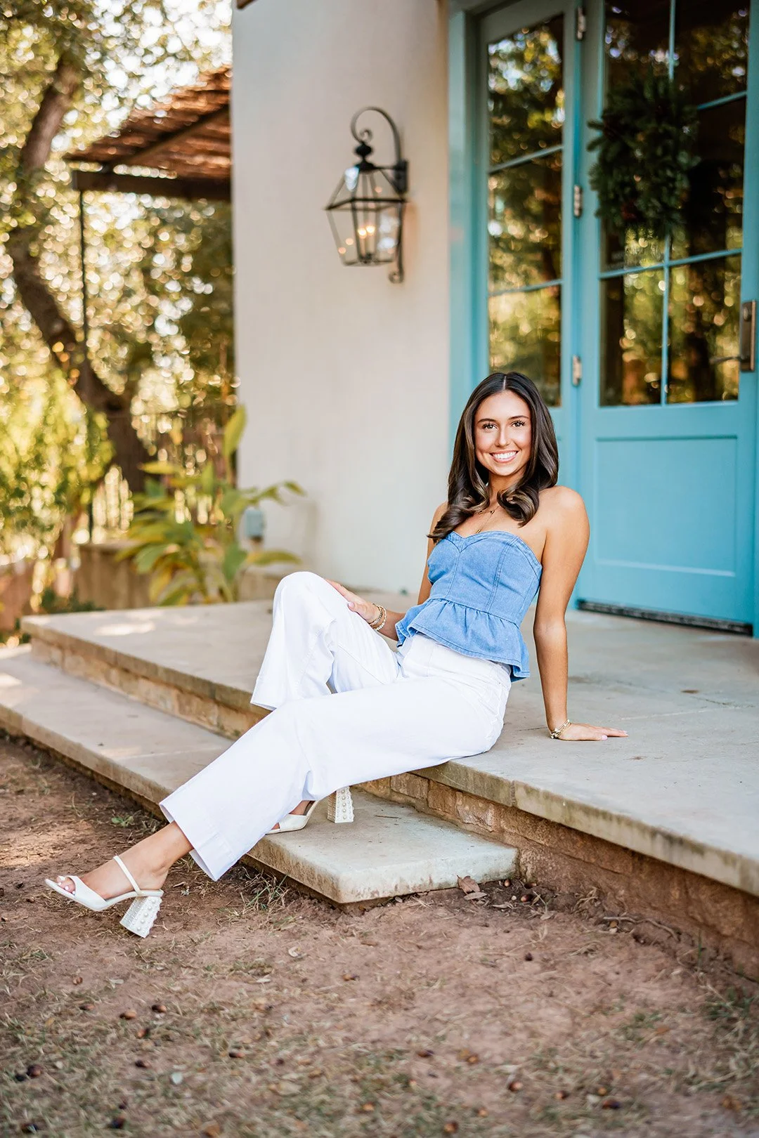 A young woman sitting on a porch steps, smiling, wearing a blue strapless top, white pants, and white high-heeled sandals, with a house with a blue door and black lantern light fixture behind her.