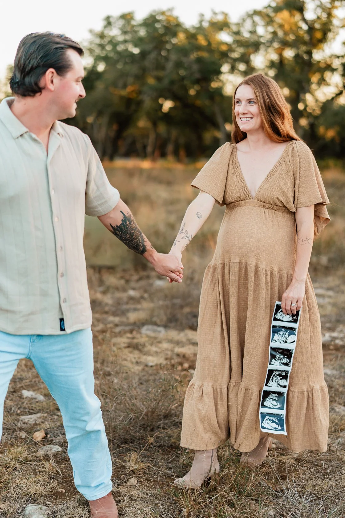 A pregnant woman in a tan dress holding ultrasound photos, holding hands with a man in a light shirt and jeans outdoors.