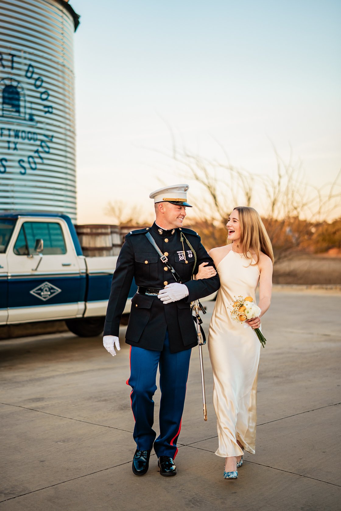 A soldier in uniform walking arm-in-arm with a woman in a cream-colored dress and holding a bouquet, both smiling, outdoors during sunset.