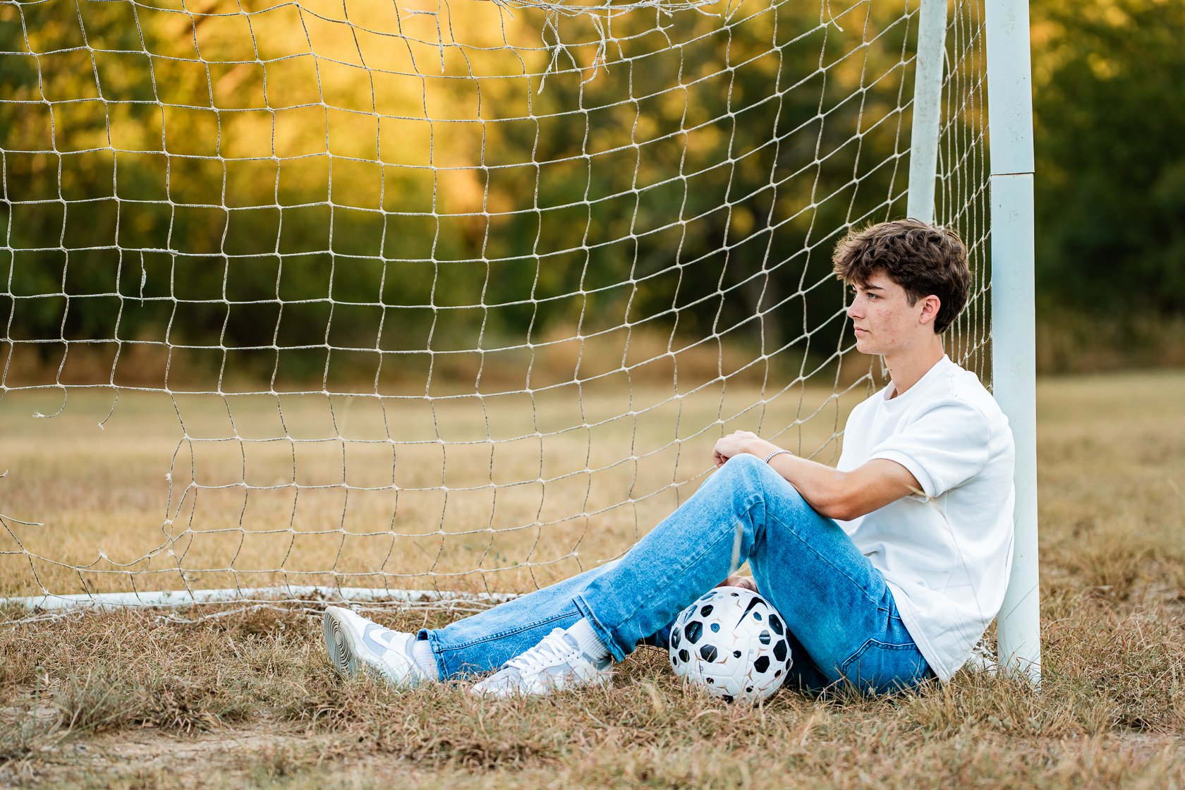 A young man sitting on the grass next to a soccer goalpost, with his back leaning against the post, a soccer ball at his feet, and a contemplative expression.