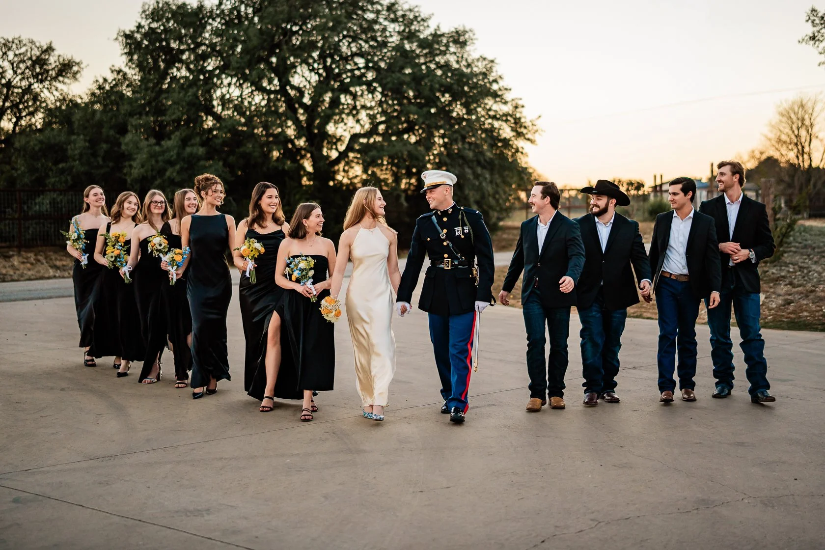 A wedding group walking outdoors at sunset, including bridesmaids in black dresses, a bride in a white gown, and groomsmen in suits with one in a military uniform, smiling and holding bouquets.