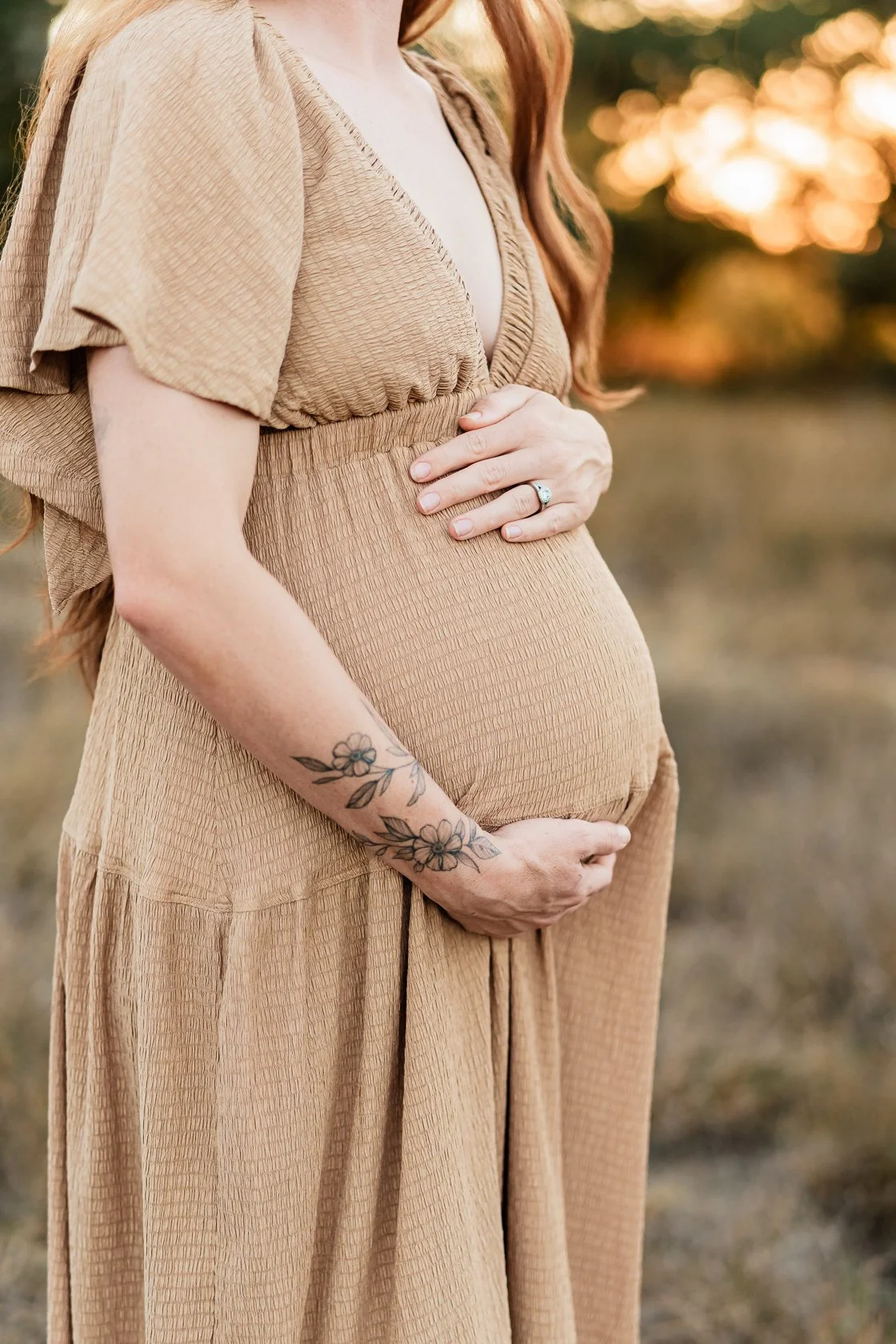 A pregnant woman gently holding her belly with both hands, standing outdoors during sunset, wearing a beige textured dress, with a floral tattoo on her forearm and an engagement ring on her finger.