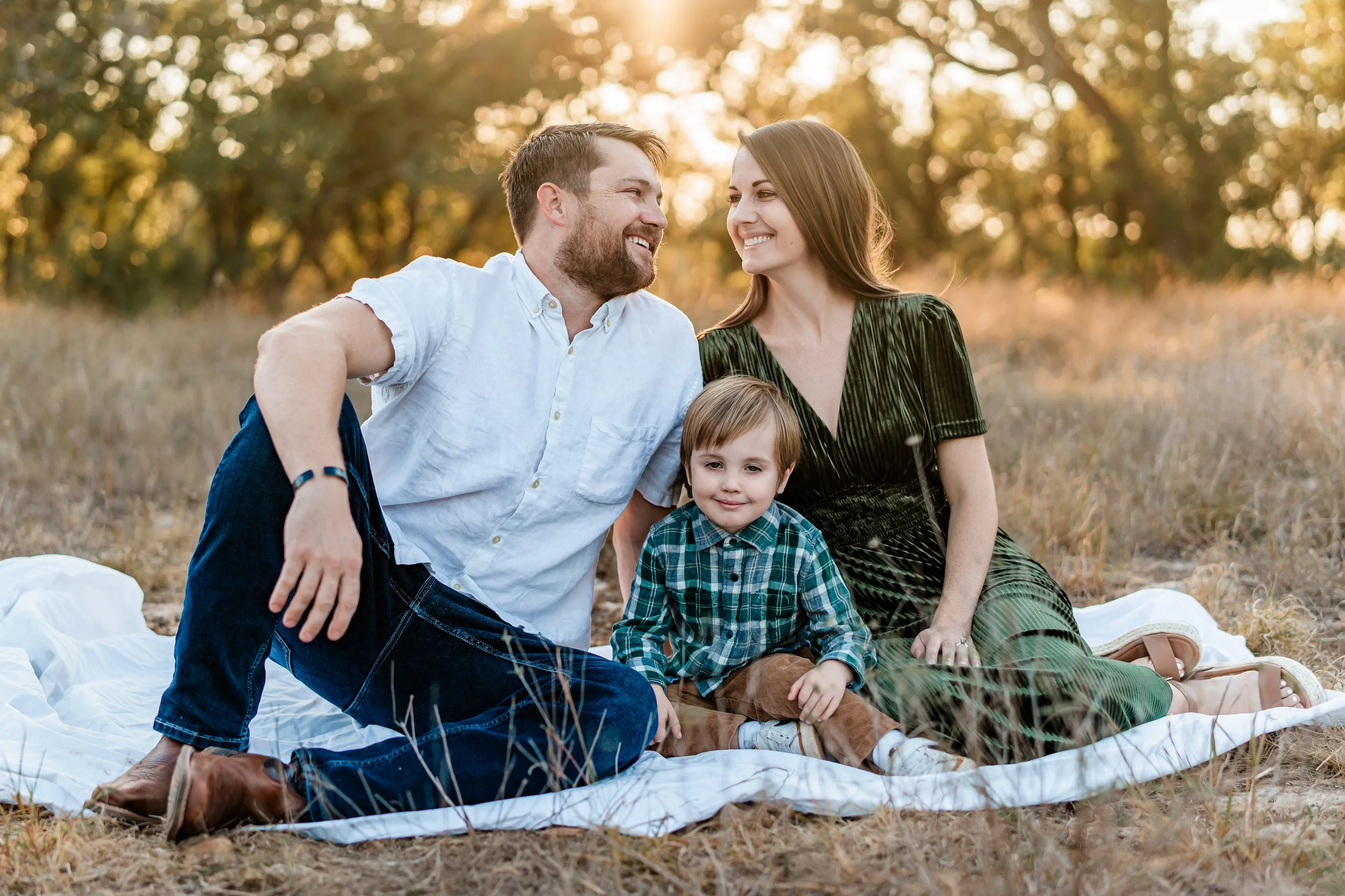 A family of three sitting on a blanket outdoors during sunset, smiling and enjoying each other's company.