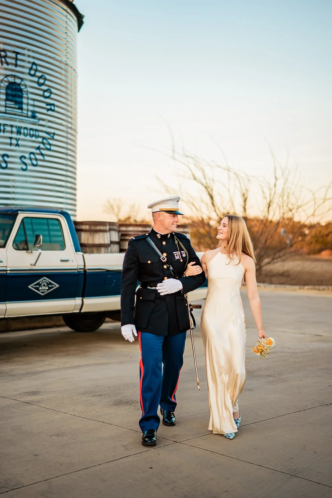 A bride walking arm-in-arm with a uniformed Marine in a desert landscape during sunset, holding a small bouquet of flowers, with a large water tower and a pickup truck in the background.
