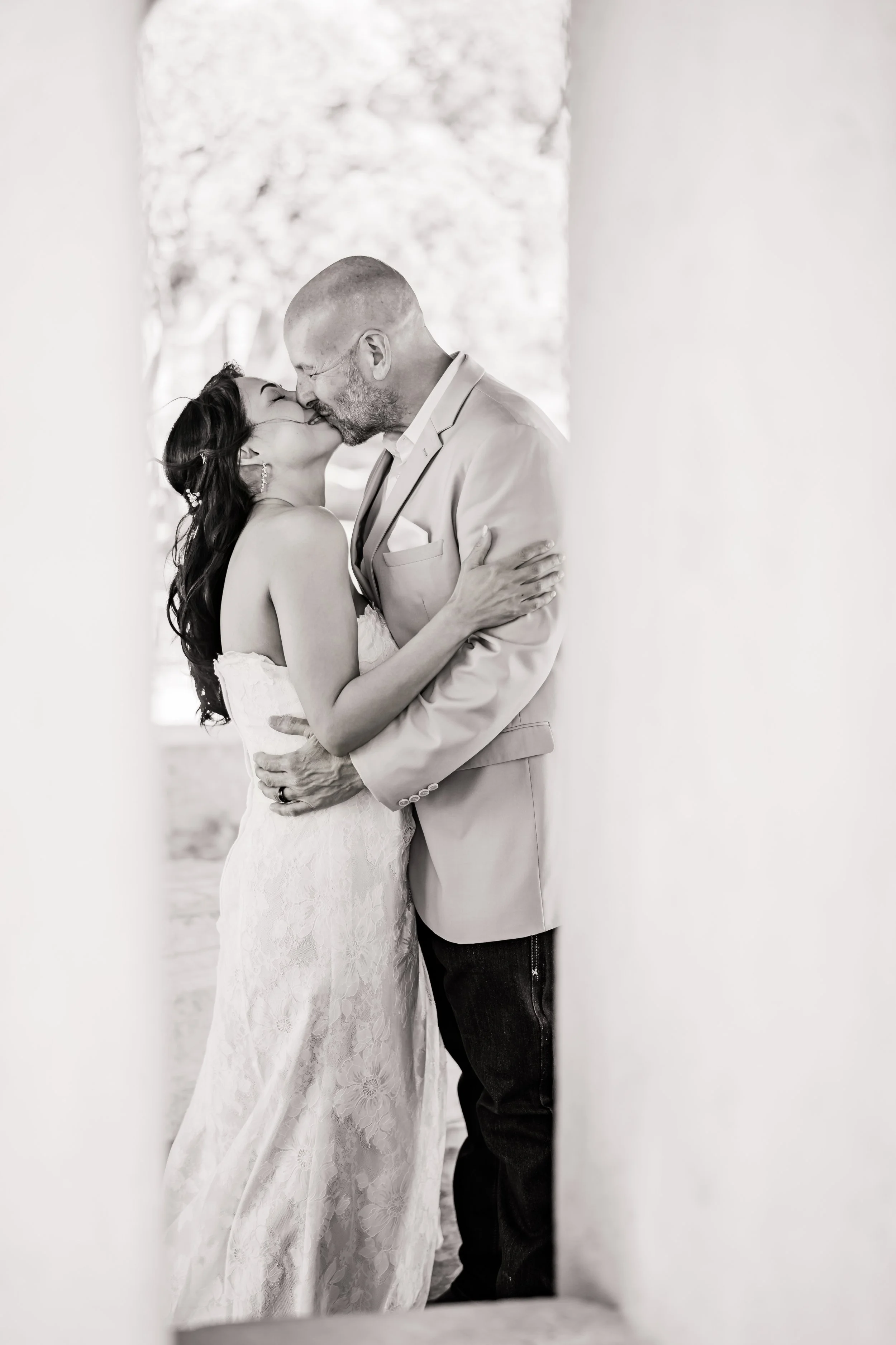 A black-and-white photograph of a bride and groom kissing, framed by two blurred vertical elements in the foreground. The bride has long dark hair and is wearing a strapless wedding dress, while the groom is bald with a beard, wearing a light-colored