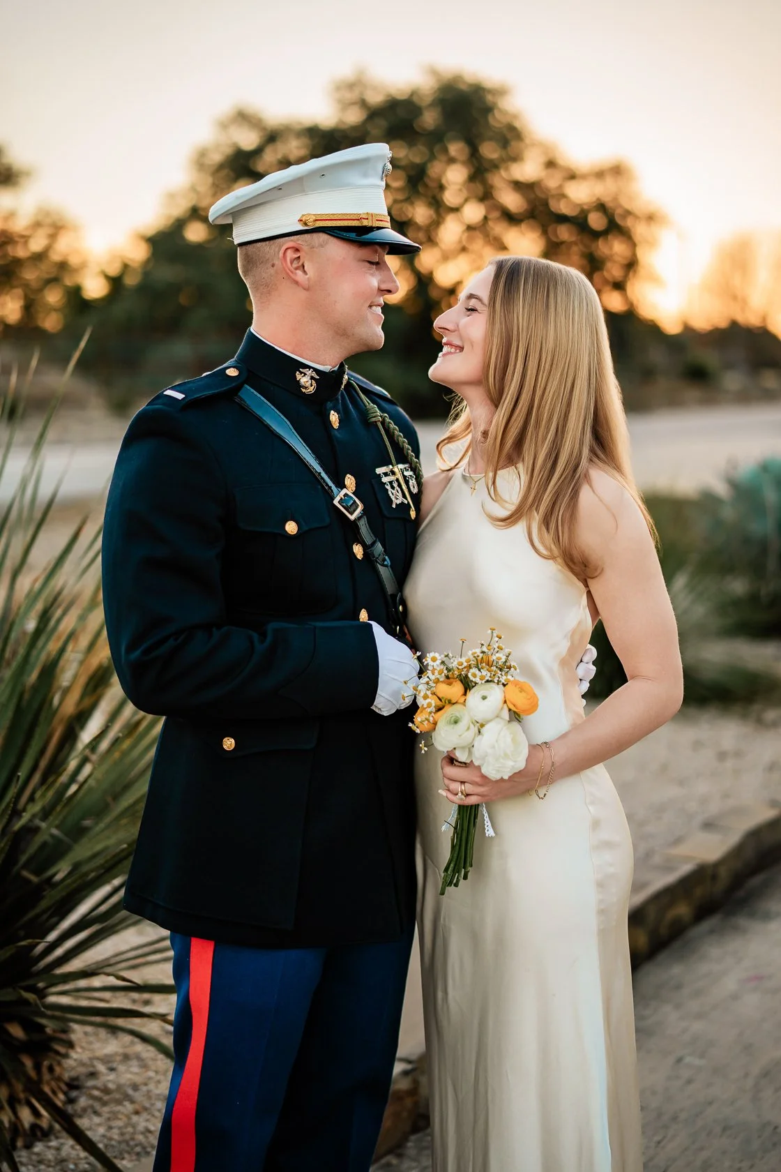 A smiling bride holding a bouquet of white and orange flowers, standing close to a groom in a United States military uniform during sunset.