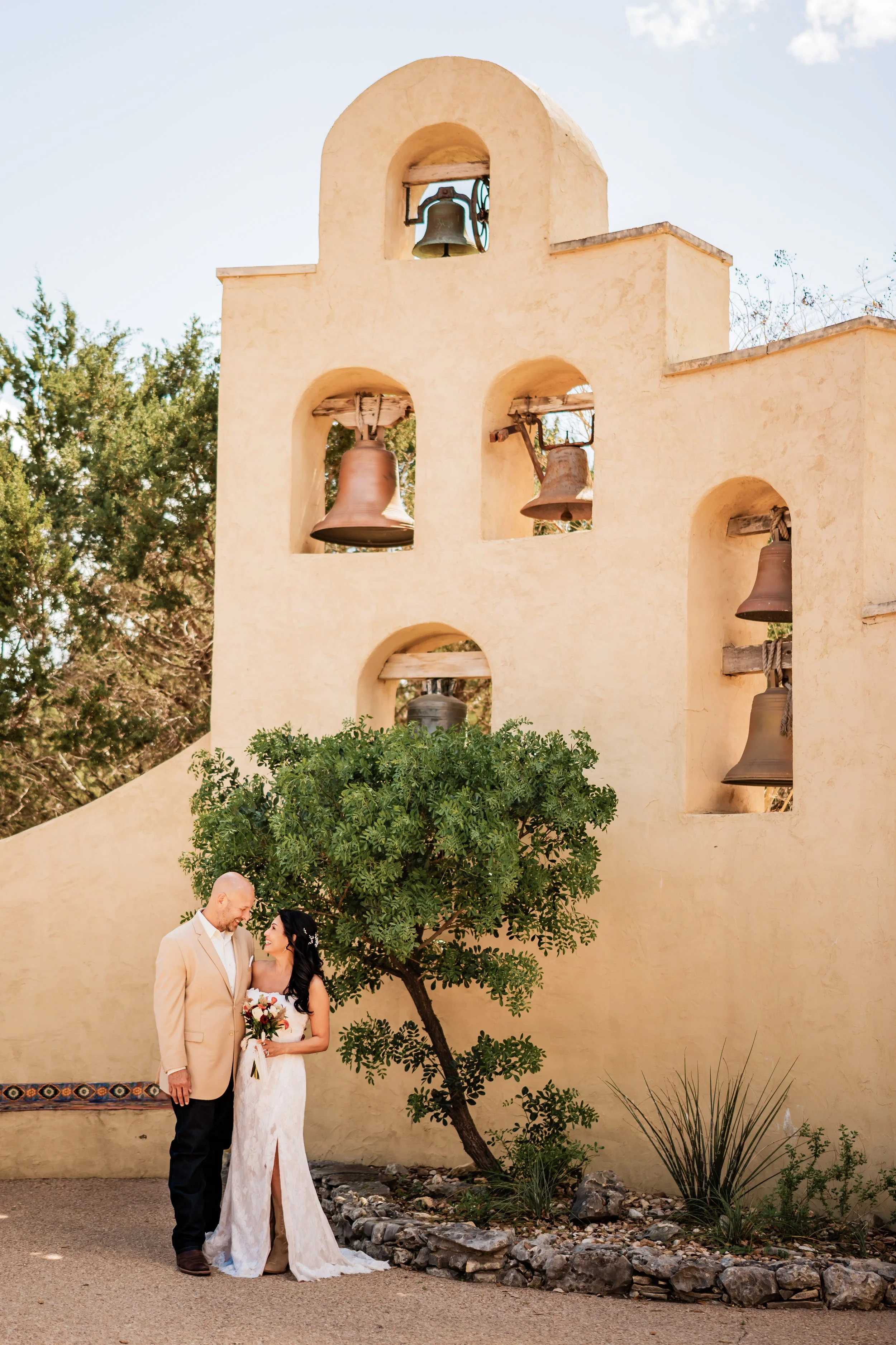 A bride and groom standing close together under a small tree in front of a beige stucco wall with bells, with greenery and rocks at their feet, on a sunny day.