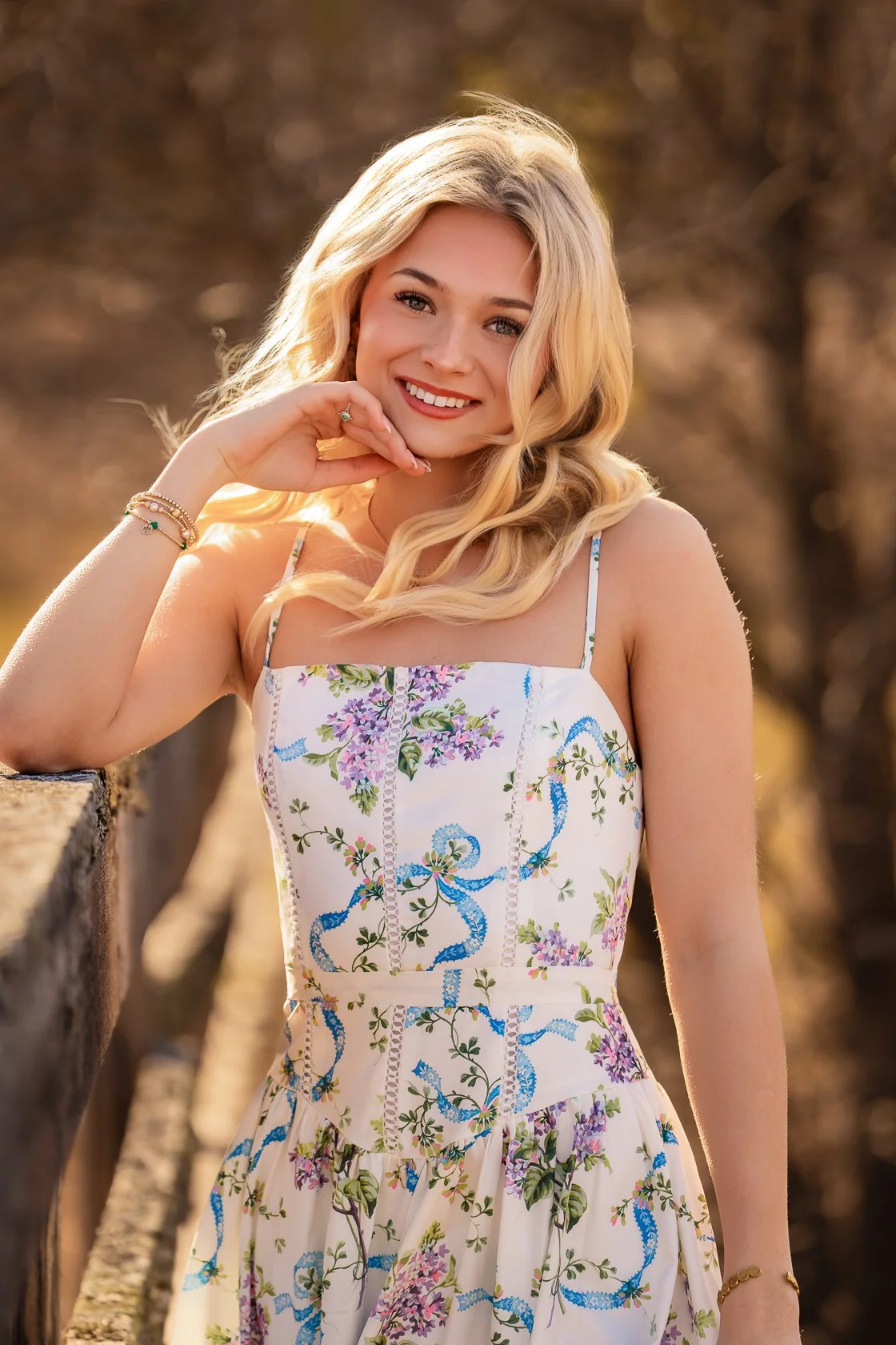 A young woman with long blonde hair smiling outdoors, wearing a sleeveless floral dress with purple, blue, and green patterns.