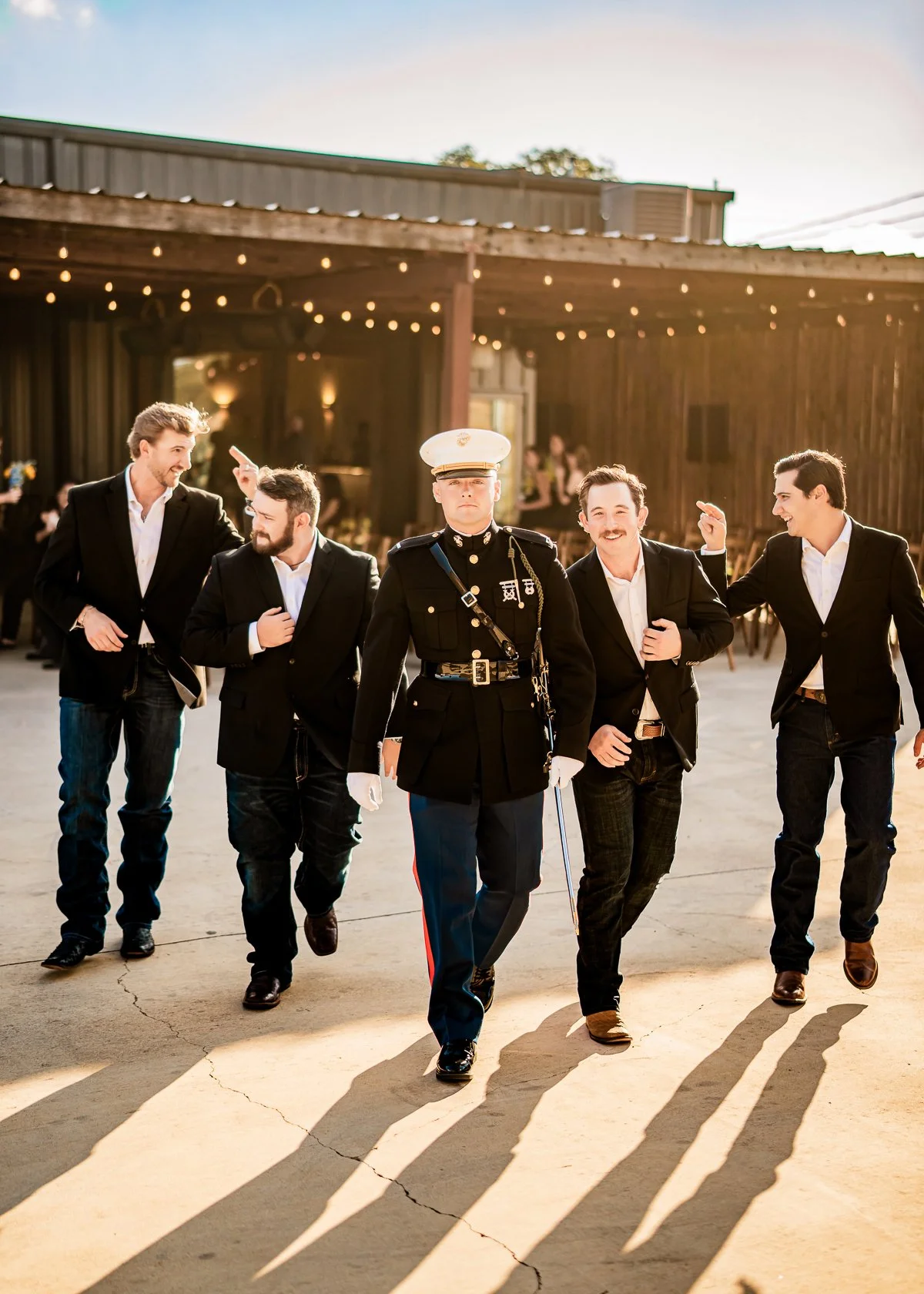 A man in a military uniform leading a group of five men dressed in tuxedos at an outdoor event during late afternoon or early evening.