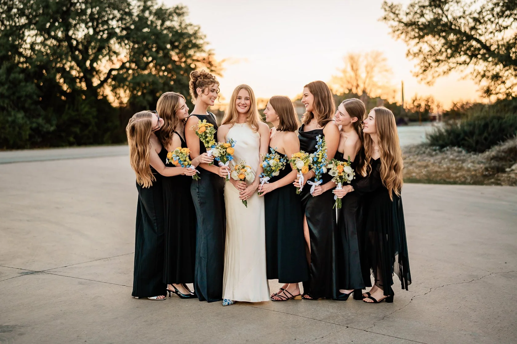 A bride in a white dress standing with seven women, all holding bouquets, outdoors during sunset.