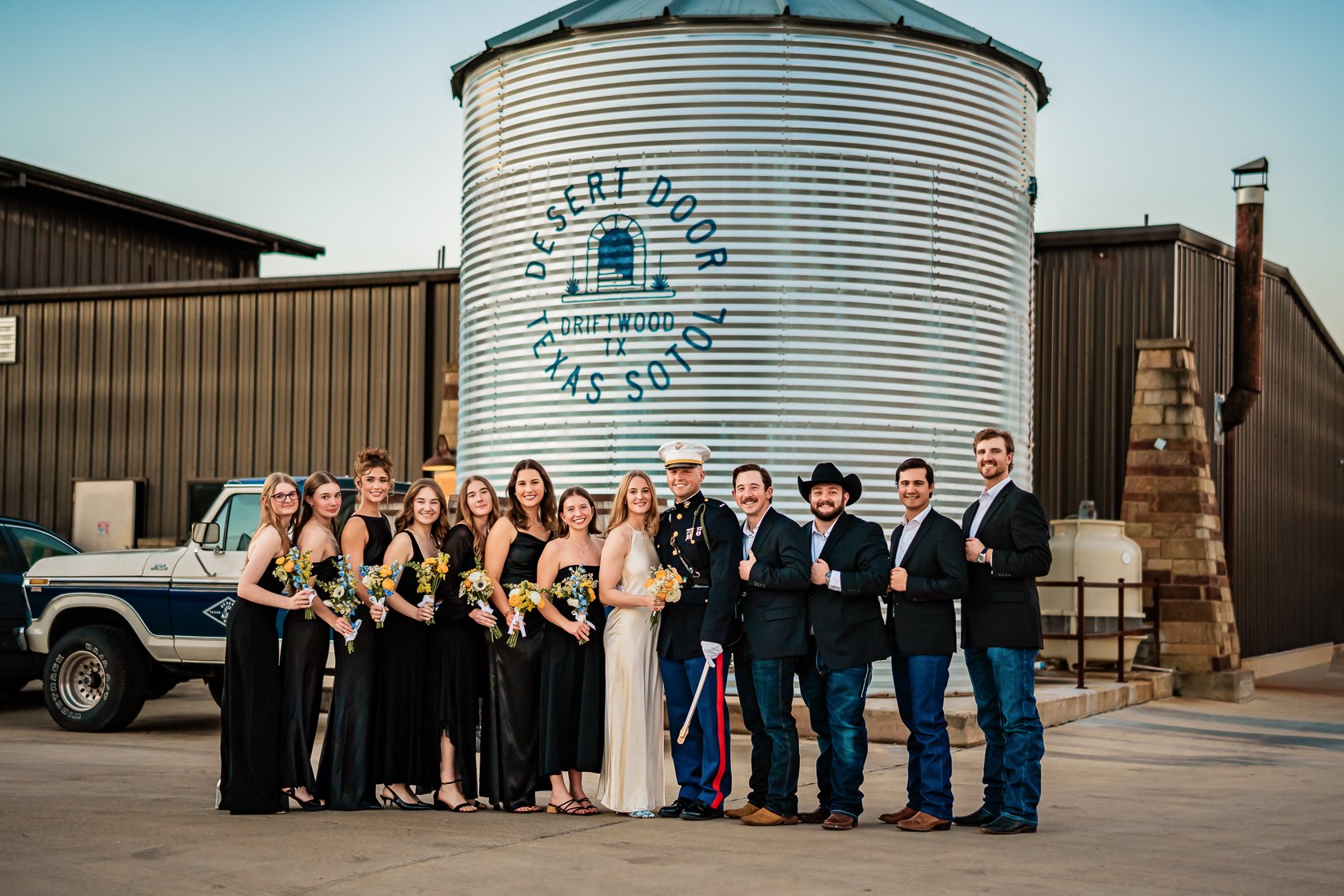 Group of people dressed in wedding attire standing outdoors in front of a large metal water tower with the text 'Desert Door Tejas Spirits' painted on it.