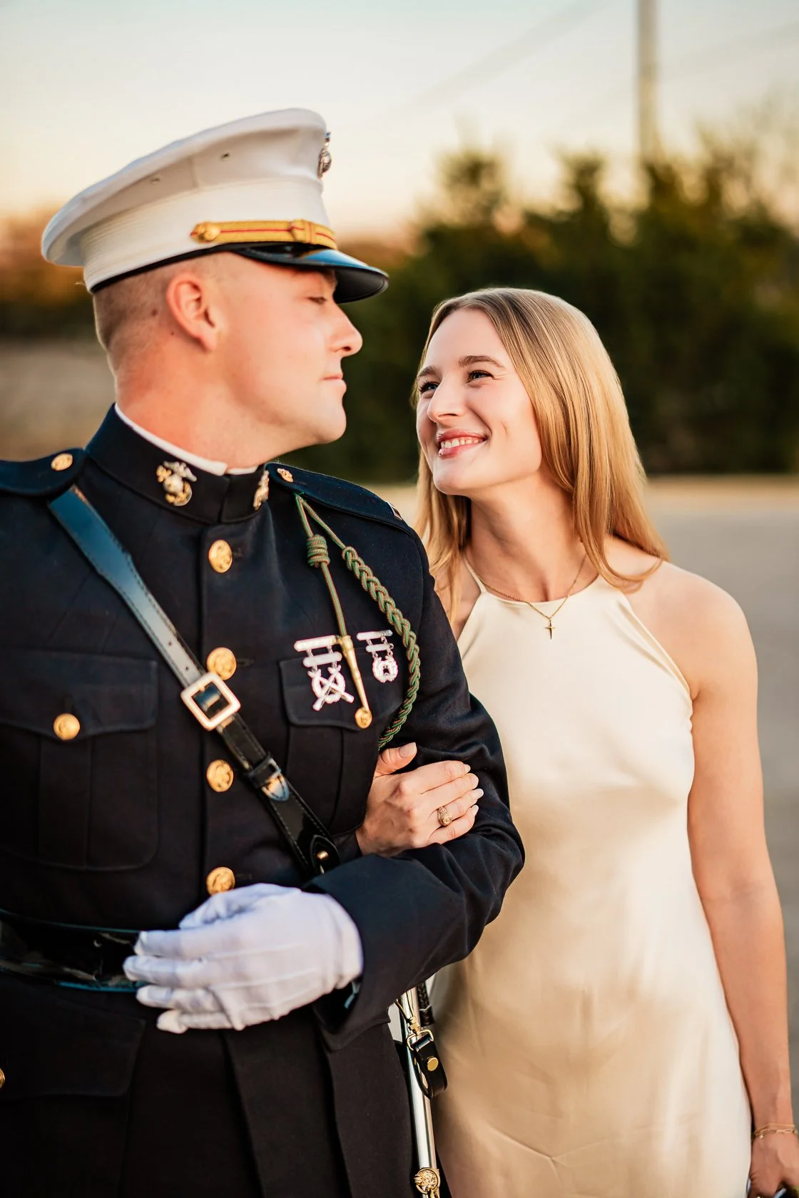 A man in a military uniform holding a woman's arm, with the woman smiling and looking at him, outdoors during sunset.