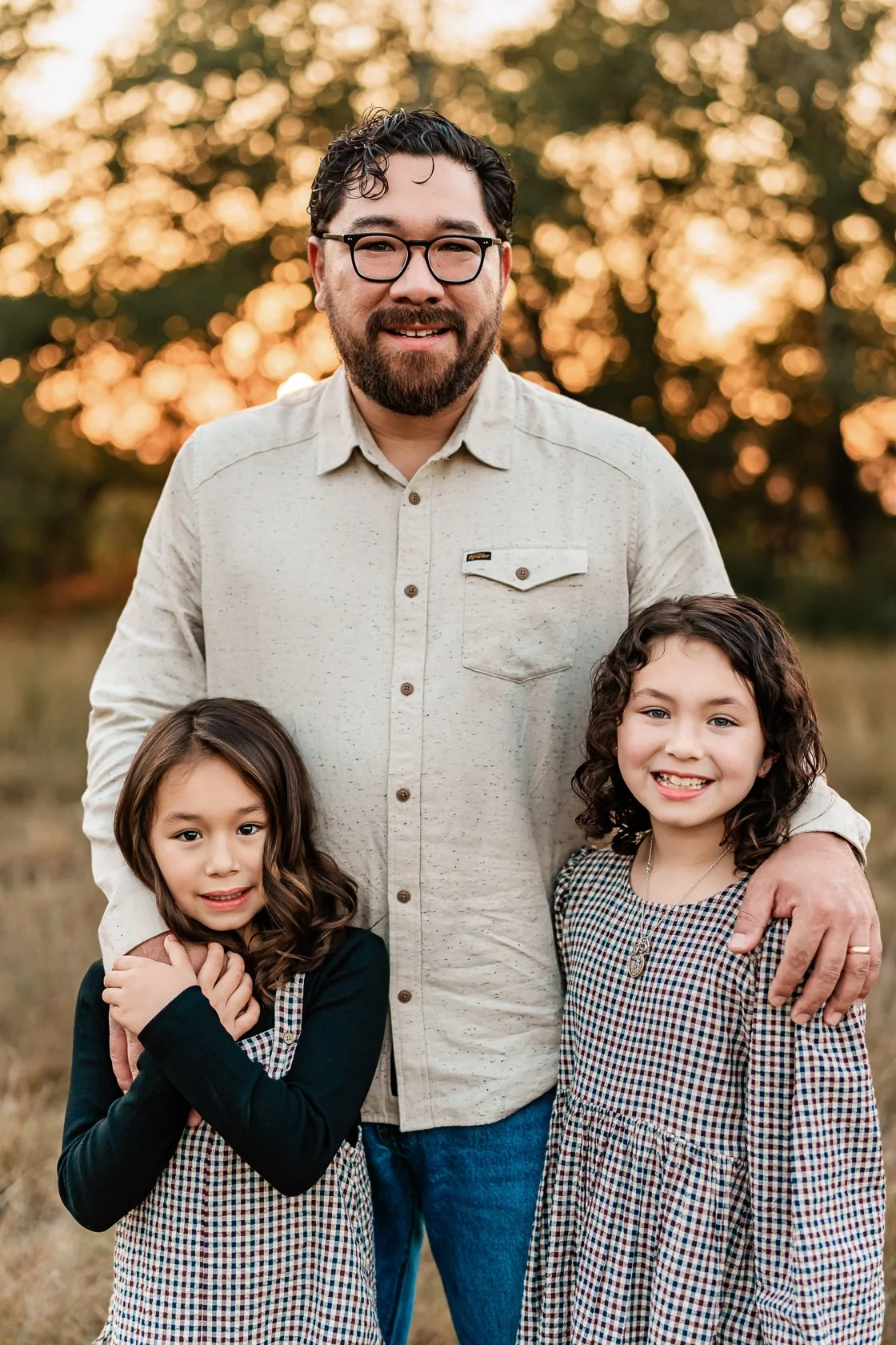 A man with glasses and a beard standing outdoors with two young girls, one with long brown hair and the other with curly brown hair, at sunset.