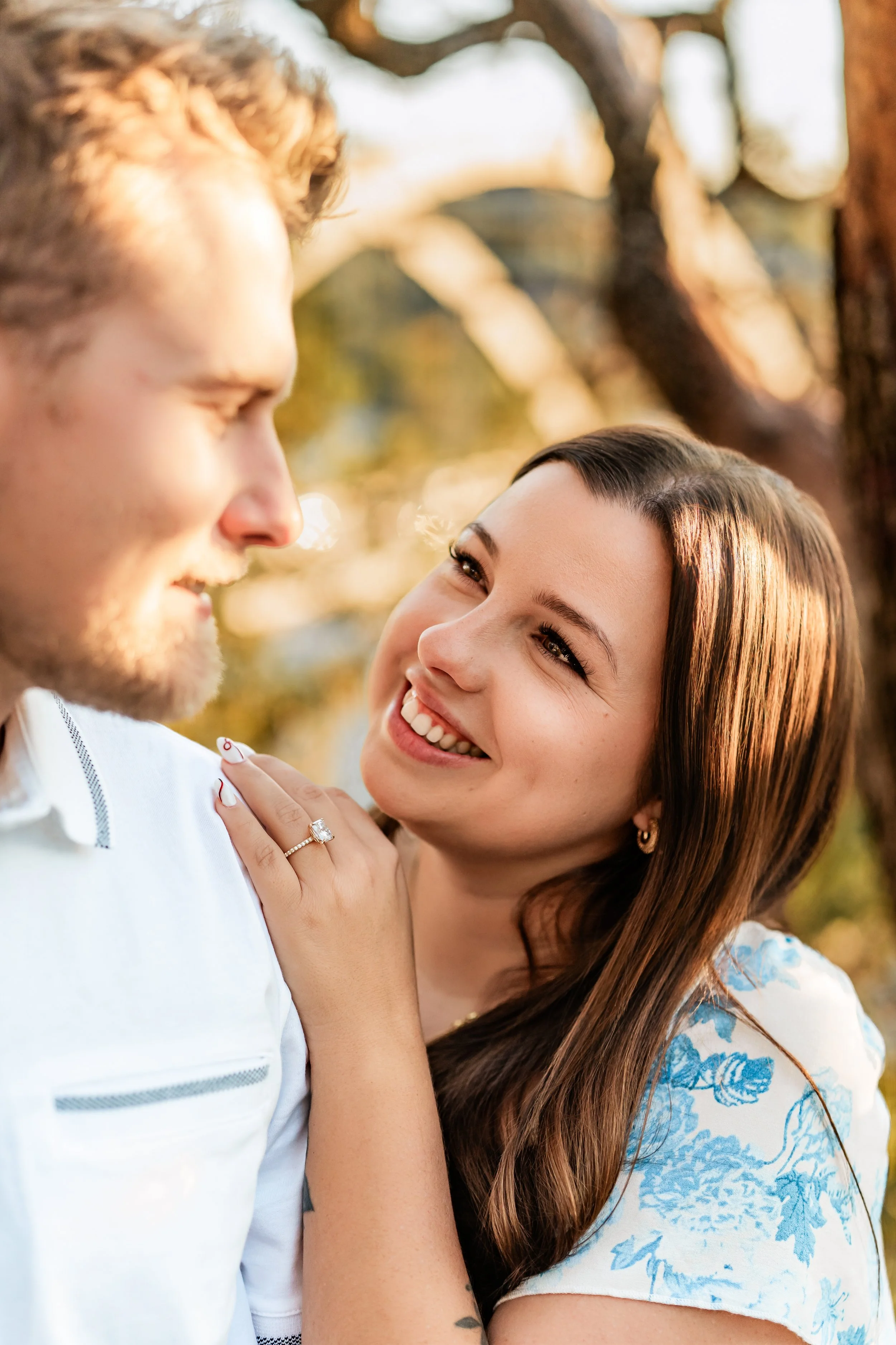 A woman smiling and touching a man's shoulder outdoors with trees and sunlight in the background.
