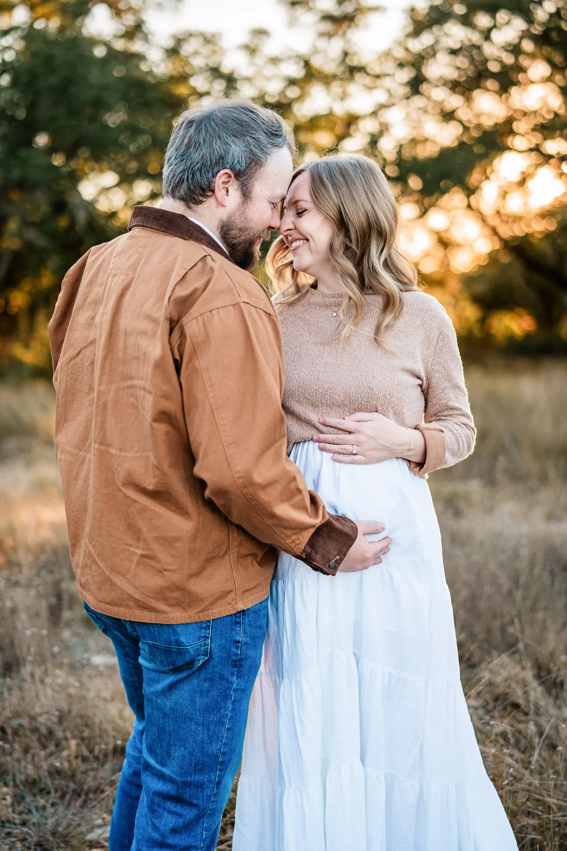 A couple standing close together outdoors during sunset, with the man touching the woman's pregnant belly and both smiling with foreheads touching.