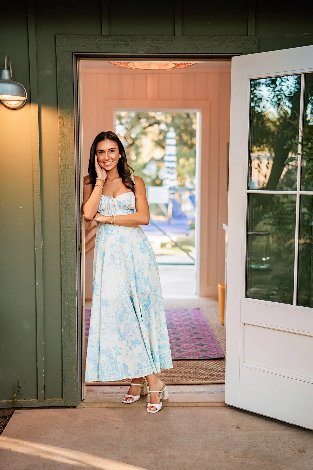 A smiling woman standing in a doorway, wearing a light blue and white floral dress with high heels, with a garden visible in the background.