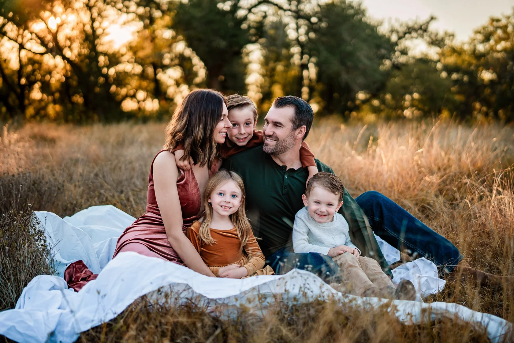 Family of five sitting on a blanket in a field during sunset, smiling and enjoying each other's company.