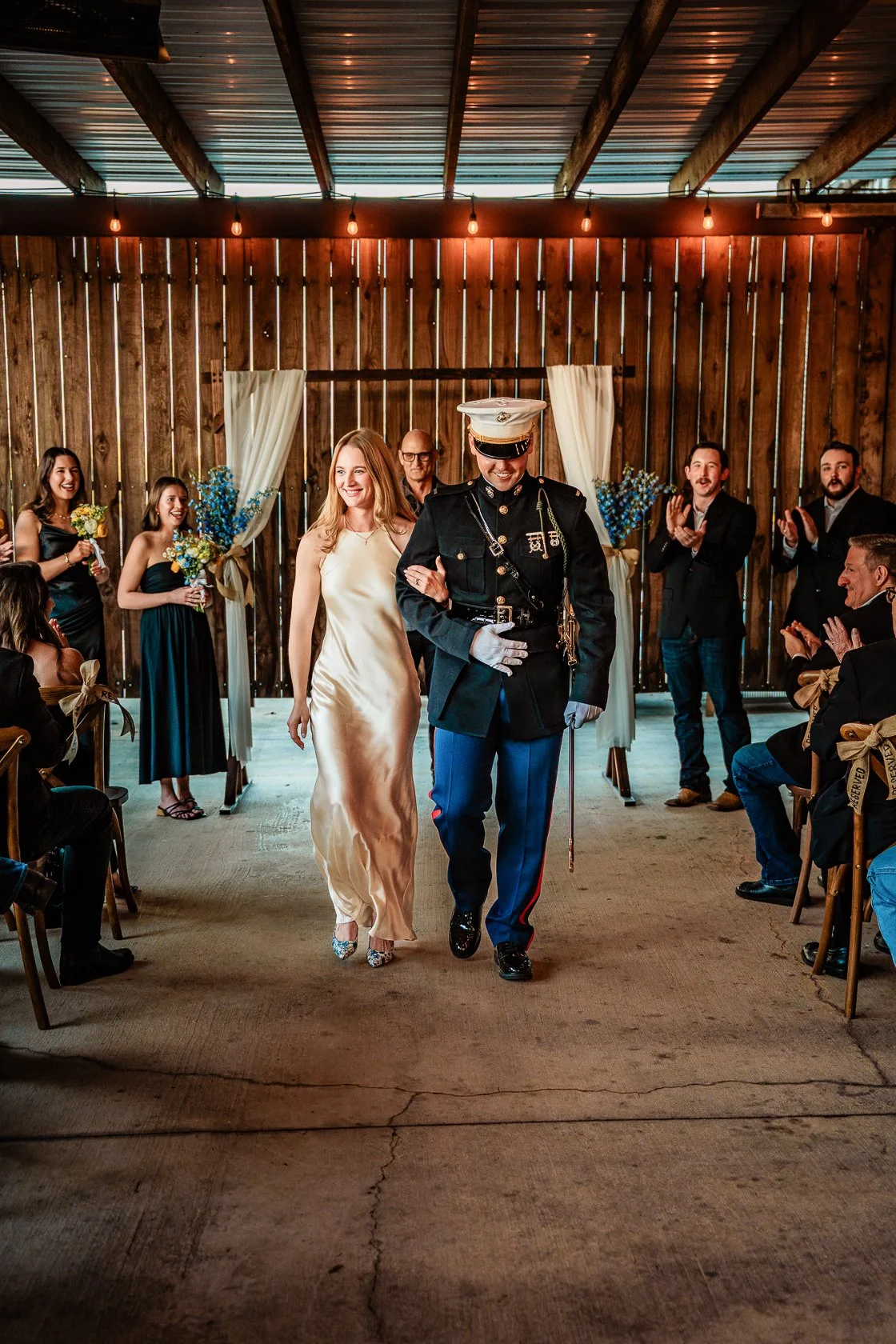A bride and a Marine walking down the aisle at a wedding ceremony with guests clapping and smiling on either side, set in a rustic wooden venue with string lights.