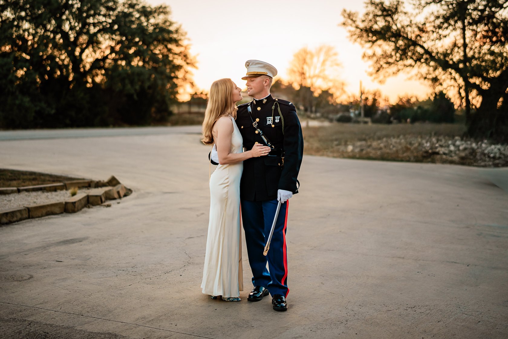 A couple standing outdoors at sunset, with the woman wearing a white dress and the man in a military uniform, close together, sharing a moment.