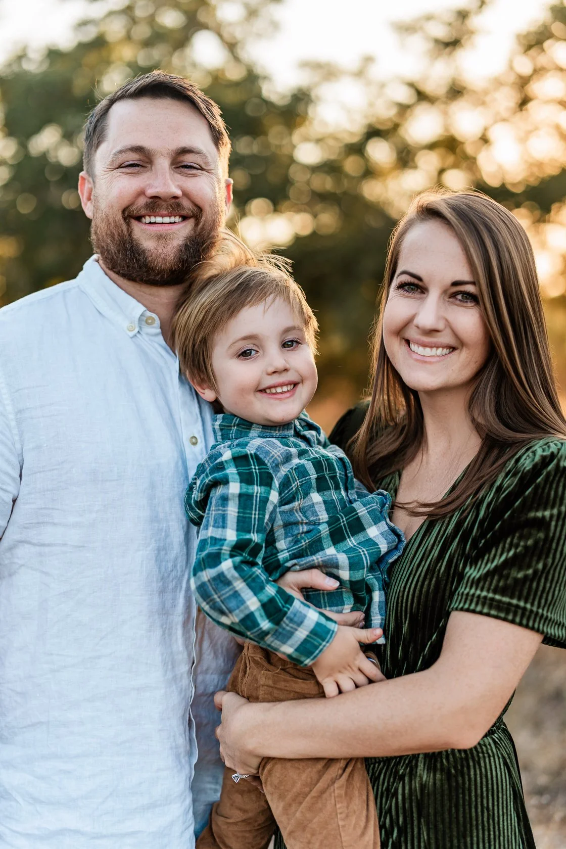 A family of three smiling outdoors during sunset, with the mother holding a young boy and the father standing beside them.