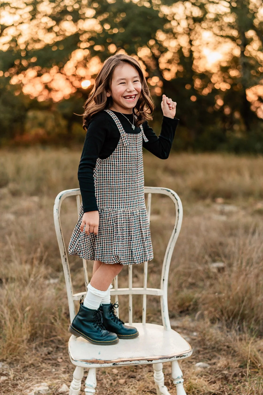 A young girl smiling and laughing, standing on an old white chair outdoors at sunset, wearing a plaid dress, black boots, and knee-high white socks.