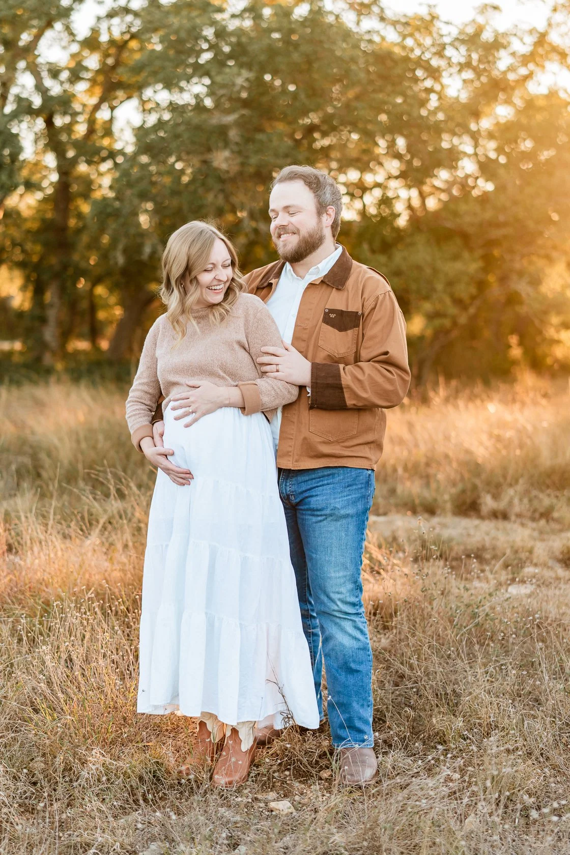 A couple standing outdoors in a field during sunset, with the woman pregnant. They are smiling and embracing each other, with trees in the background.