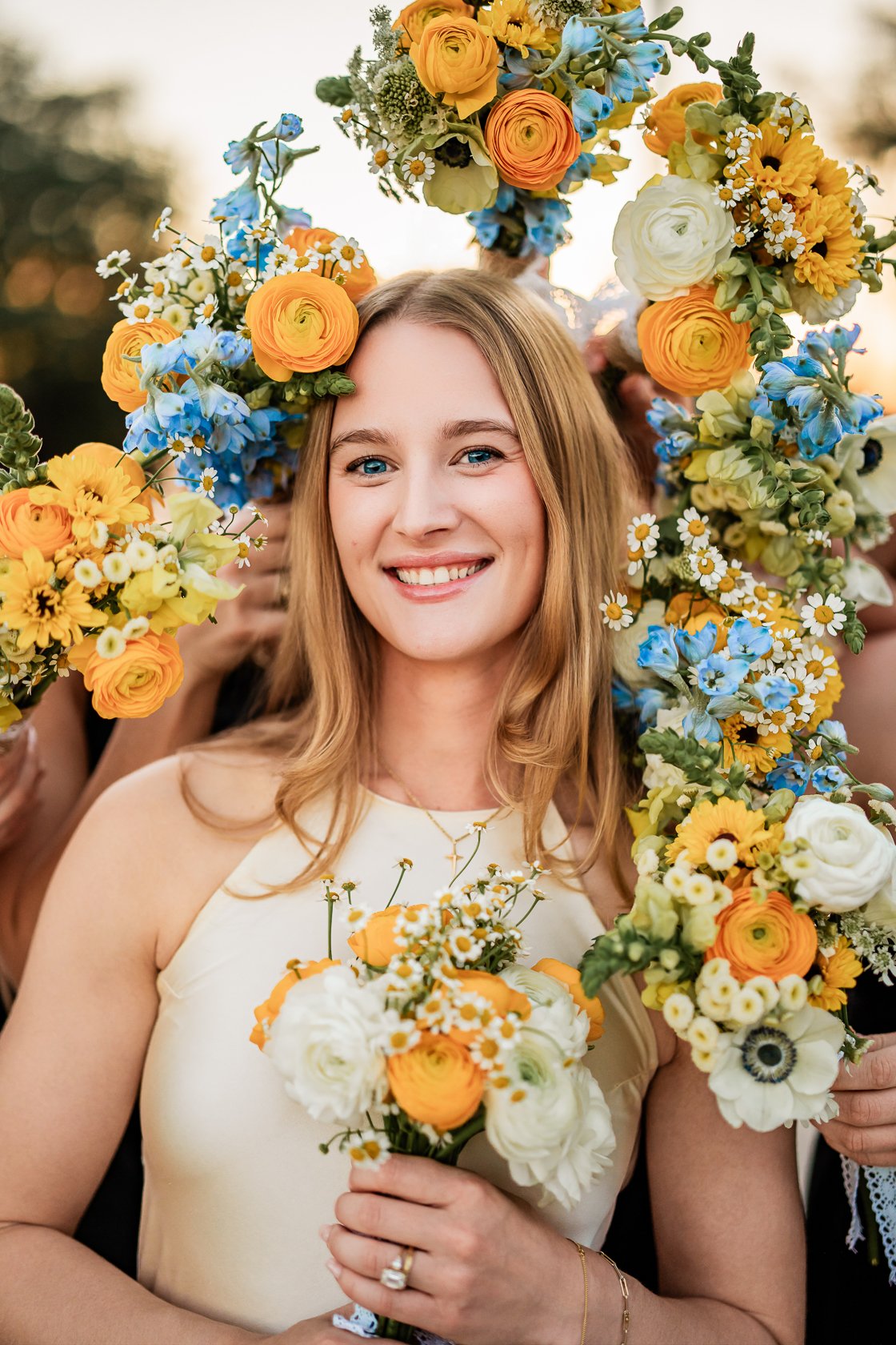 A smiling woman with blonde hair holding a bouquet of yellow, orange, and white flowers, surrounded by friends holding similar bouquets during a celebration.