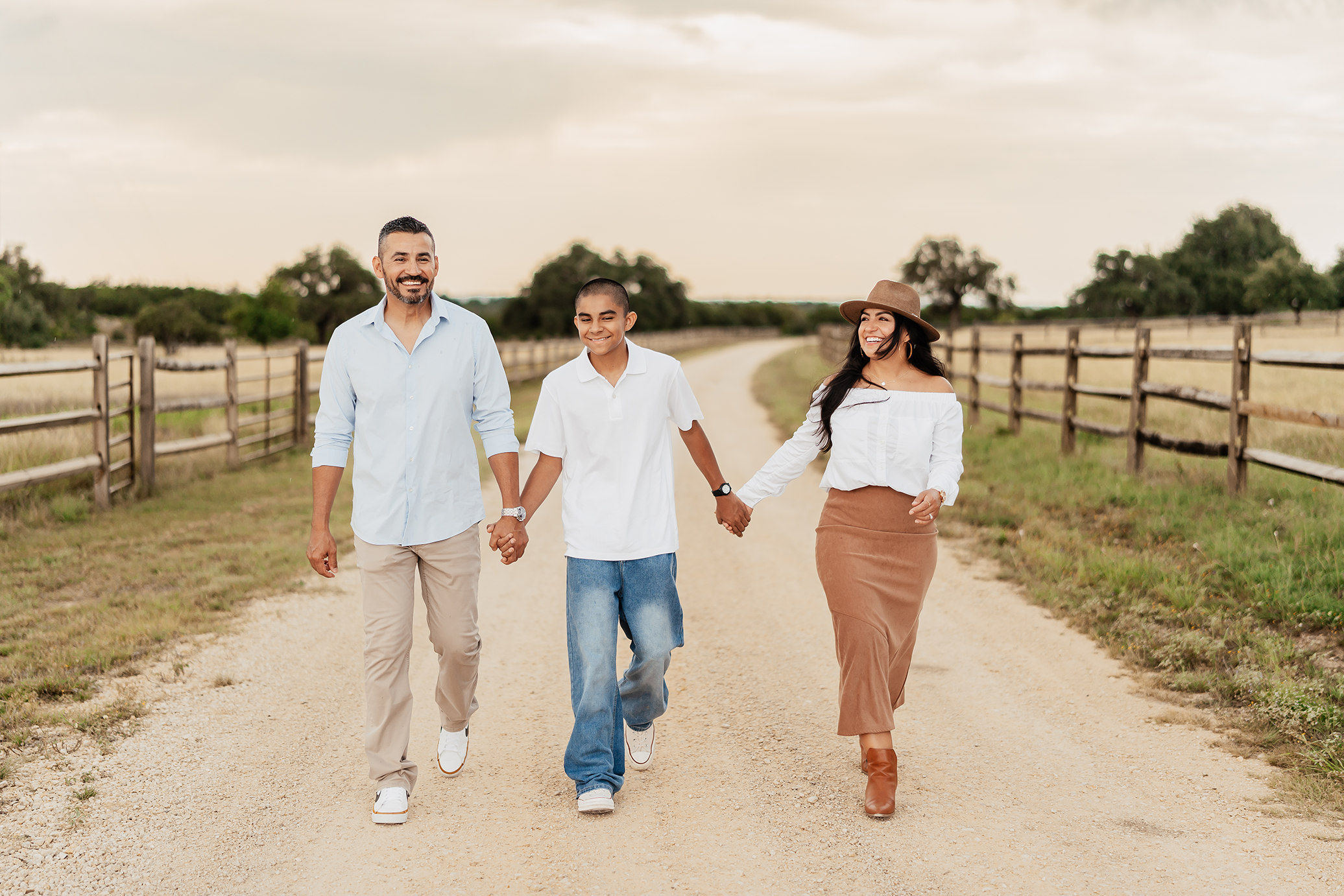 A family of three walking hand in hand on a dirt path in a rural area with wooden fences on both sides, enjoying a cloudy day.