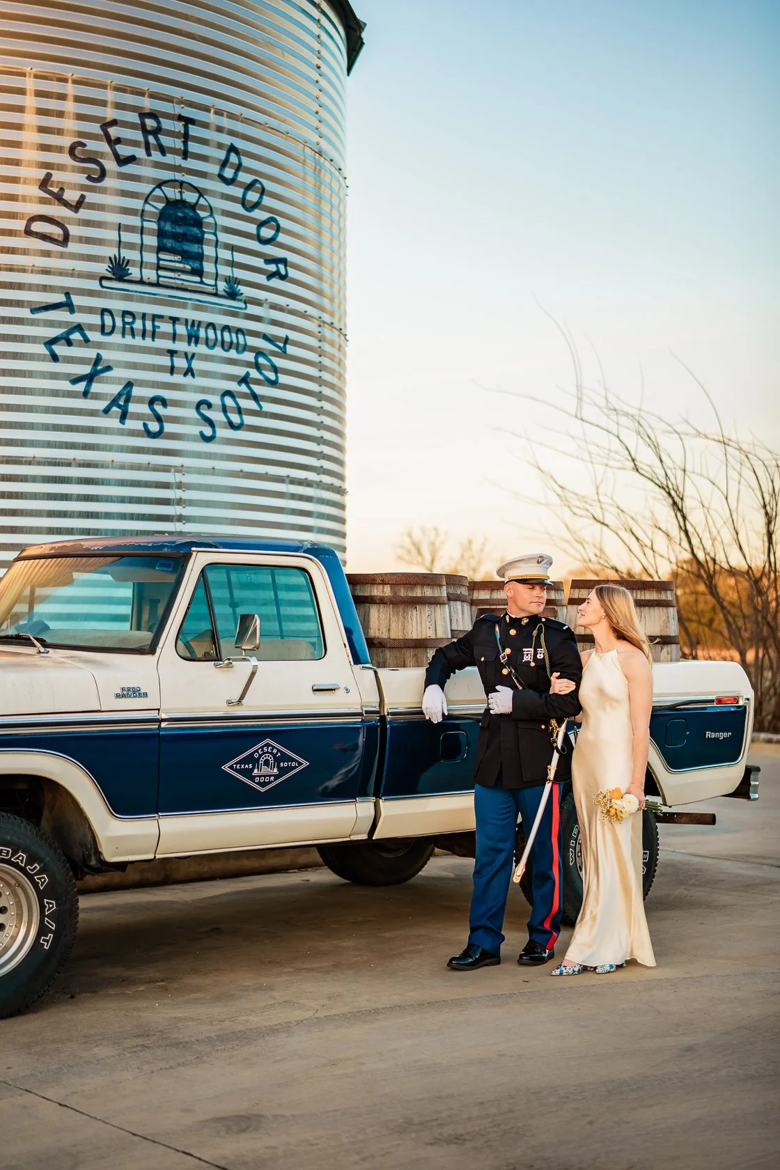 A couple in wedding attire standing next to a vintage pickup truck in front of a large water tower with Texas and Salty Texas written on it.