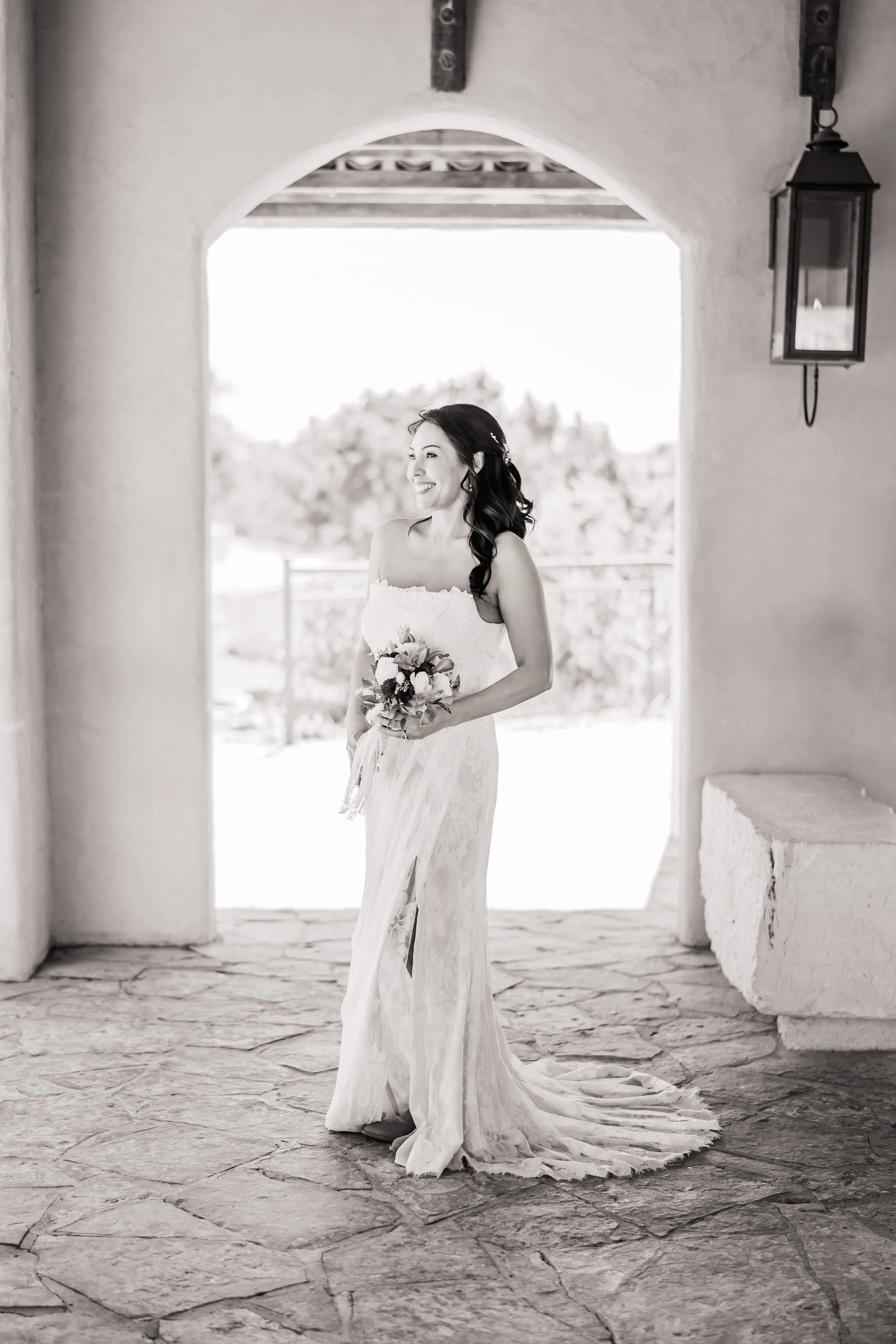 A bride in a strapless lace wedding gown holding a bouquet, standing in an arched doorway outdoors, smiling and looking to her left, in black and white.