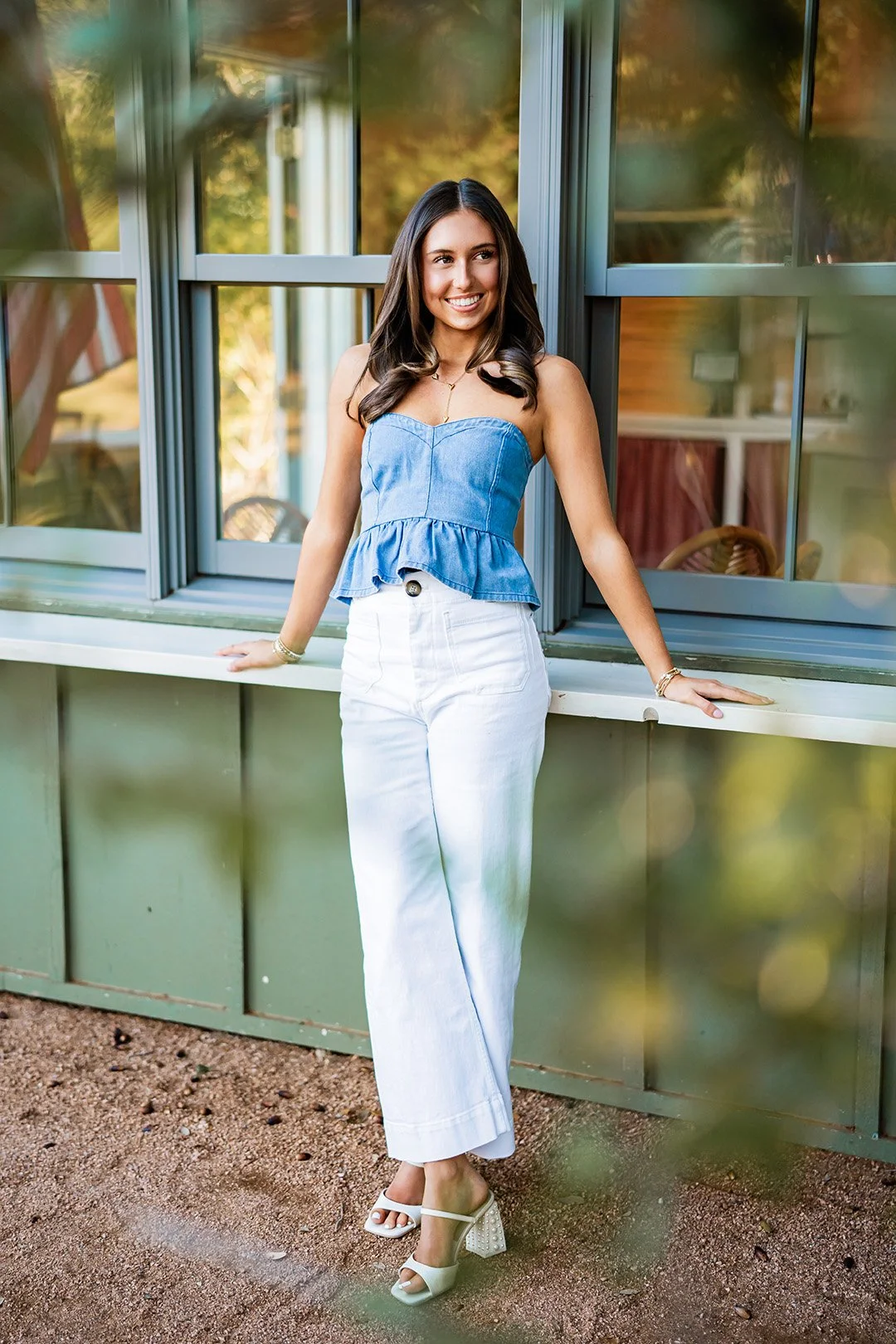 A young woman with long dark hair is standing outside in front of a green house wall and window, smiling and leaning on the windowsill. She is wearing a strapless denim top, white wide-leg pants, and white heeled sandals.