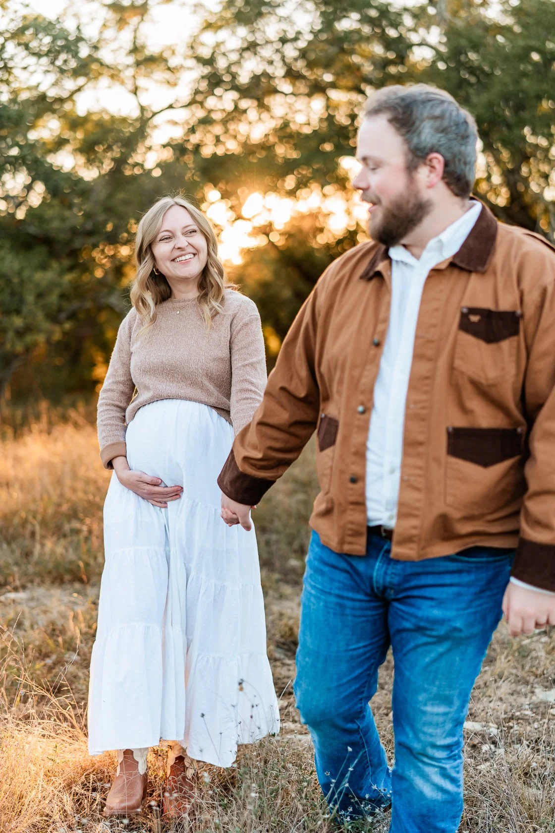 A pregnant woman in a white skirt and beige sweater holding her belly, smiling at her partner outdoors during sunset, holding hands with her partner, who is wearing a brown jacket and blue jeans, in a natural setting with trees in the background.
