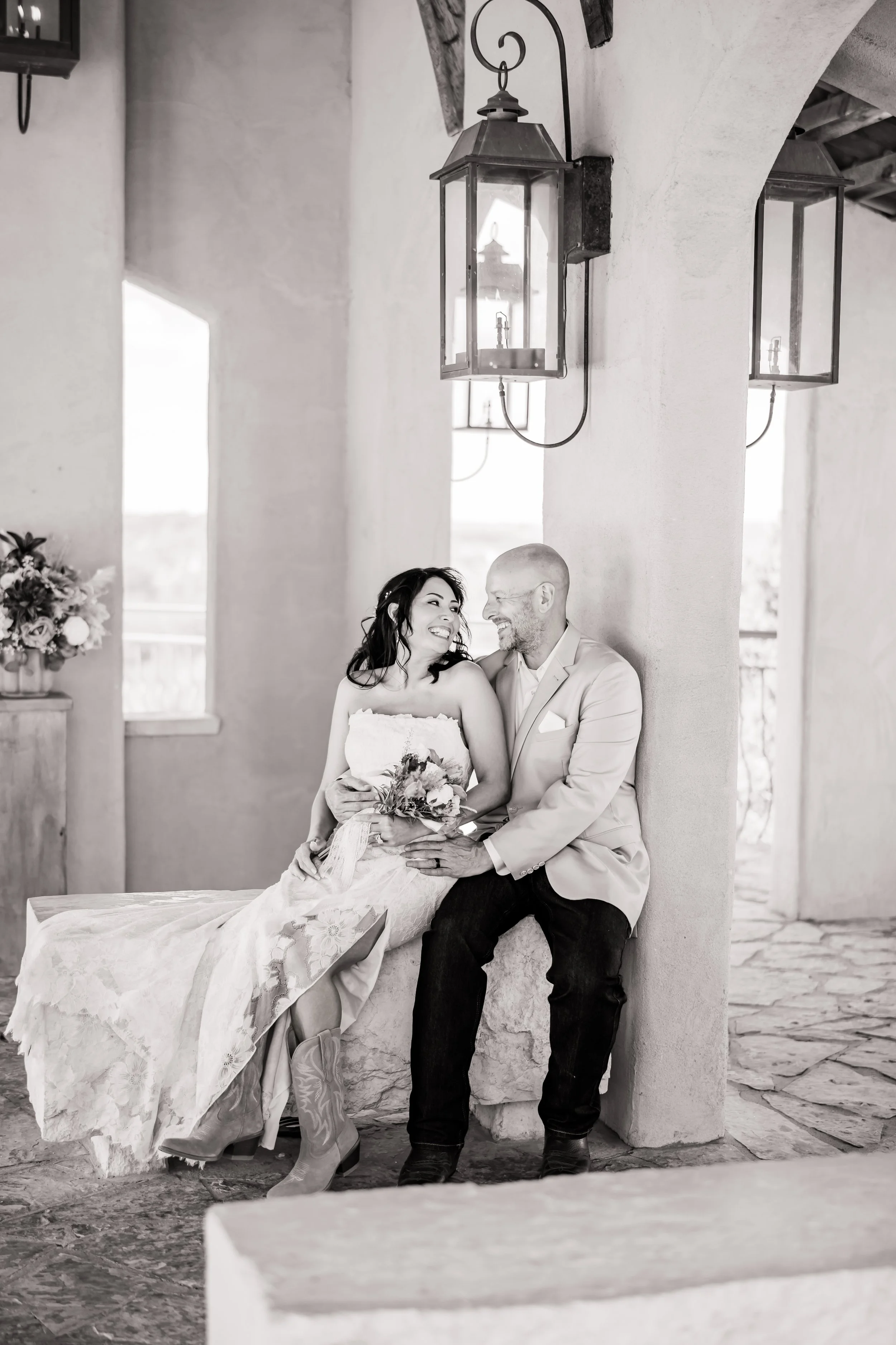 A black and white photo of a bride and groom sitting together, smiling and looking at each other, on a stone bench under hanging lanterns.