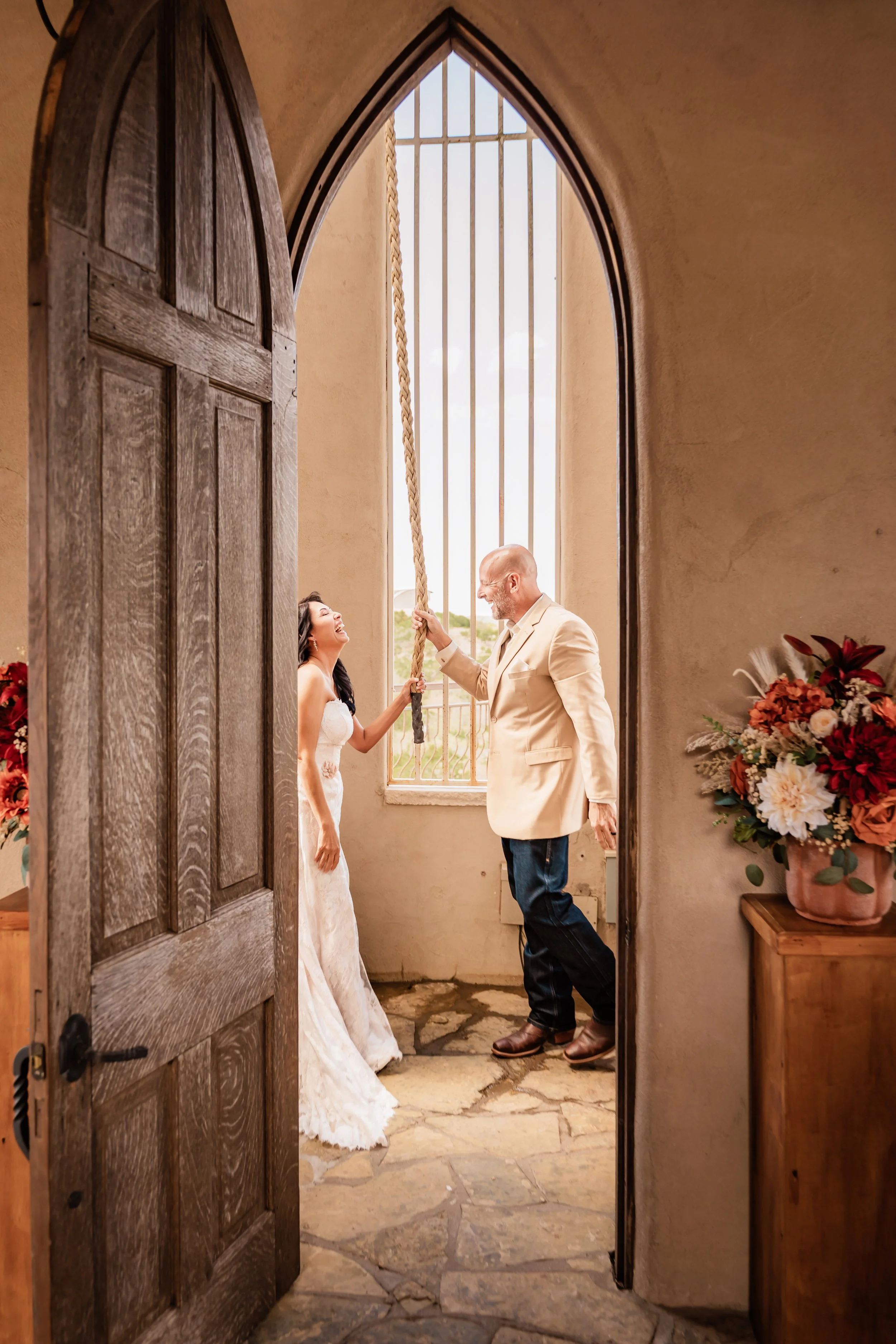 A bride and groom share a joyful moment inside a chapel with a large arched window, wooden door, and floral decorations.
