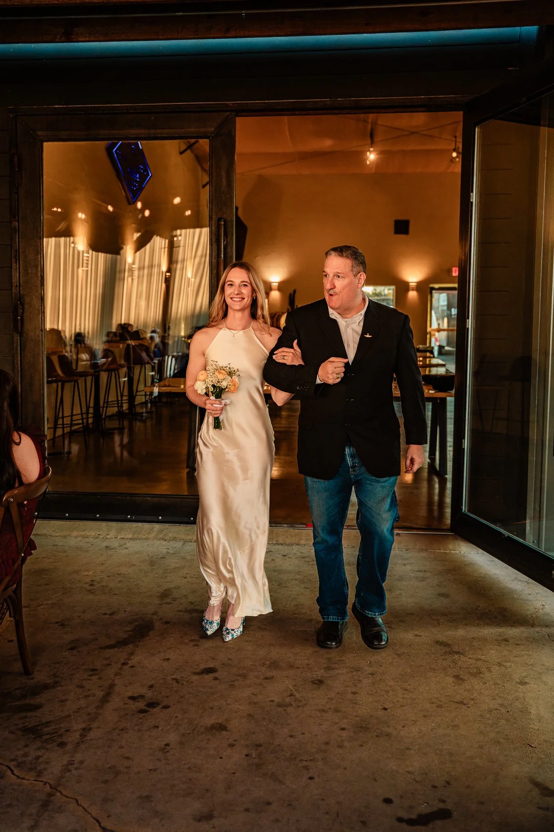 A woman in a long, satin wedding dress holds a bouquet of flowers and walks arm-in-arm with a man in a black blazer, jeans, and white shirt through a doorway at a wedding reception. They are smiling and appear happy.